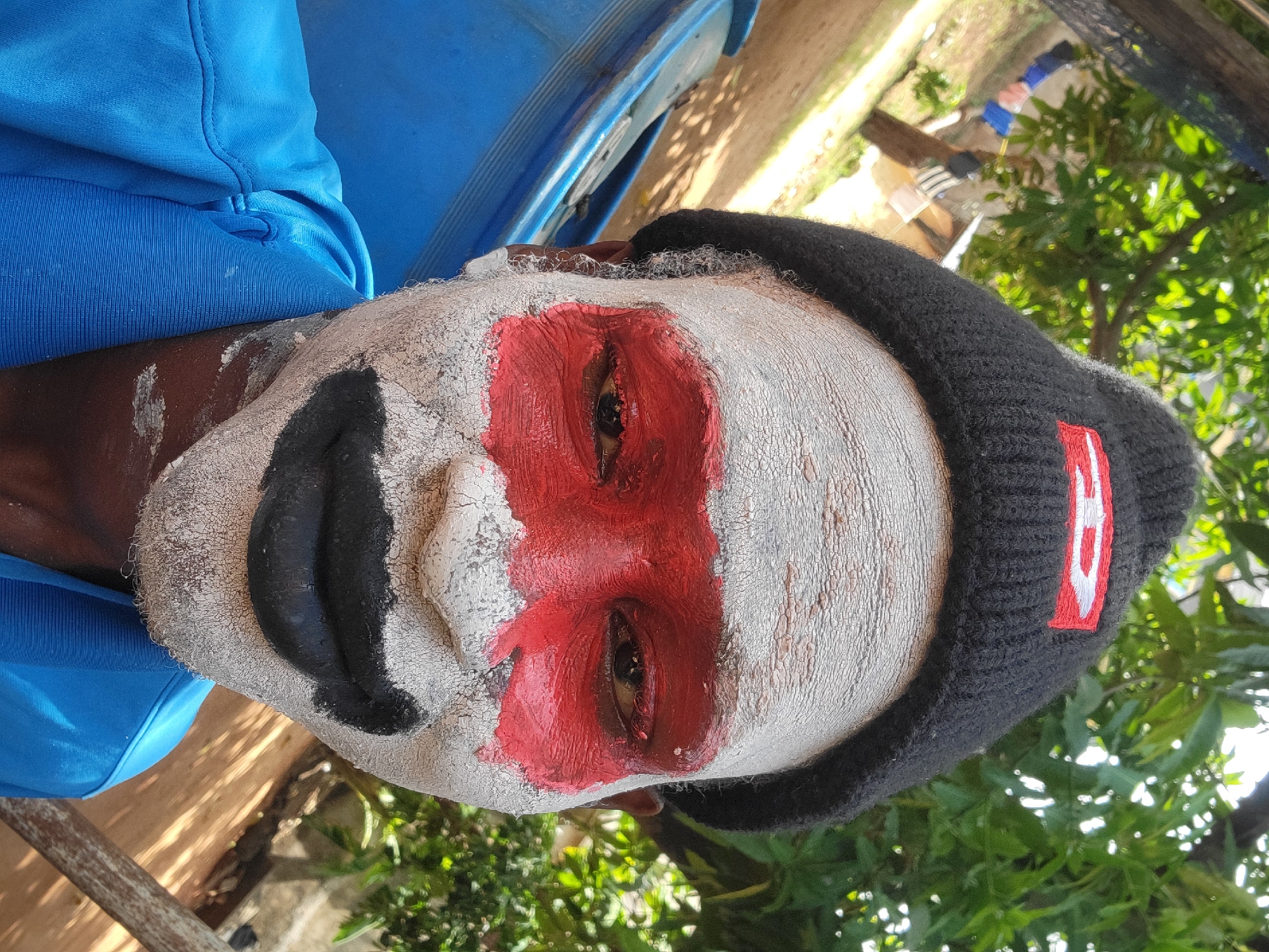 A person with white face paint, a red stripe painted across the eyes, and black paint on the nose is wearing a black beanie with a red and white stripe. The person is wearing a blue shirt and is sitting next to a blue barrel. The background includes trees and foliage, suggesting an outdoor location. The city and specific location are not identifiable.