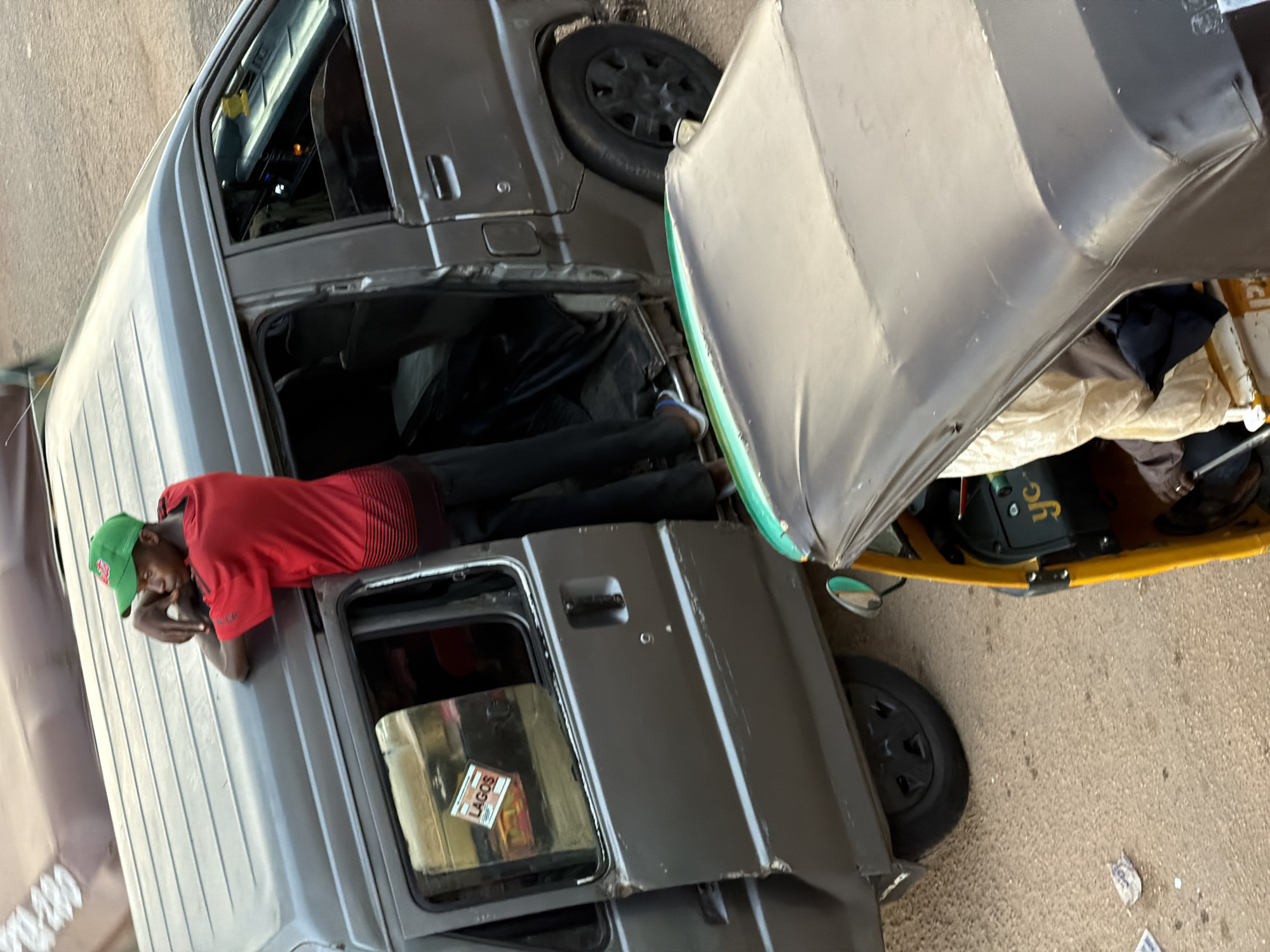 A young man wearing a green cap and a red t-shirt stands with his arms on the roof of a gray van in what appears to be a street in Bukuru, Nigeria. The van's passenger door is open, and the man is looking into the vehicle. Another vehicle, a yellow and gray auto-rickshaw, is parked in front of the van, partially obscuring it from view. The ground is a dusty, reddish-brown surface, suggesting an outdoor, urban environment. There are no other people clearly visible or interacting in the scene. The lighting suggests it is daytime.