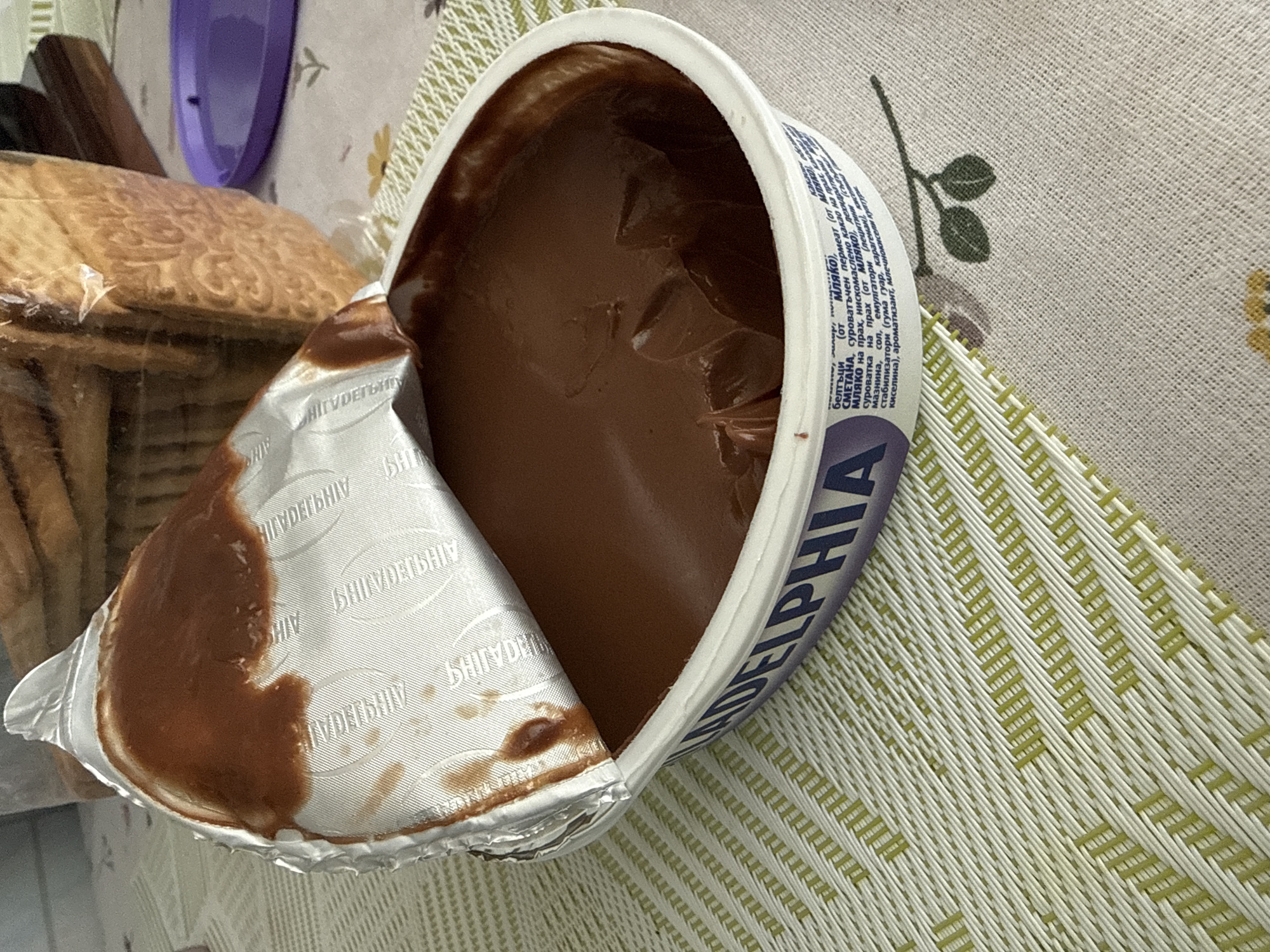 A close-up, eye-level shot shows an opened tub of chocolate cream cheese. The foil lid is peeled back, revealing the smooth, dark brown contents within. The tub itself is white with purple lettering that partially spells out "PHILADELPHIA." In the background, out of focus, is a loaf of bread wrapped in plastic and a purple lid. The tub sits on a placemat with a woven texture and a subtle floral pattern. The lighting is bright, casting soft shadows.