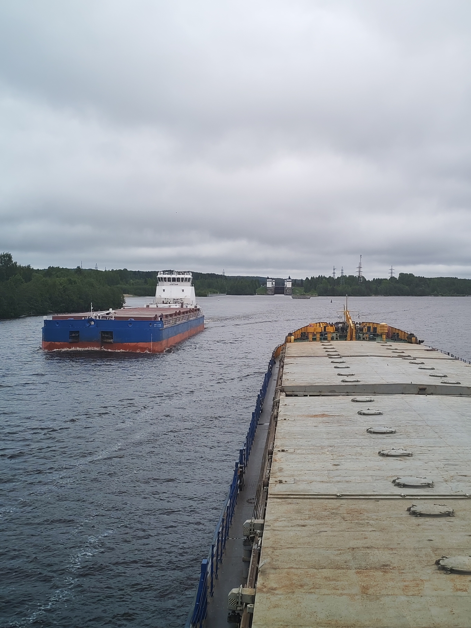 The image depicts a wide river landscape with two large barges navigating the water. In the foreground, the deck of a barge is visible, with a faded gray surface featuring numerous access hatches. Yellow structures are visible near the rear of the barge. To the left, another barge is moving in the opposite direction. It is painted with a red and blue hull and has a white, multi-story structure at the top. In the distance, a structure with a tall white tower is visible, possibly a lock or dam. The landscape includes green trees lining the riverbanks and utility towers visible on the shoreline. The sky is overcast with heavy clouds. The location is in the city of Анхимово, Russia.