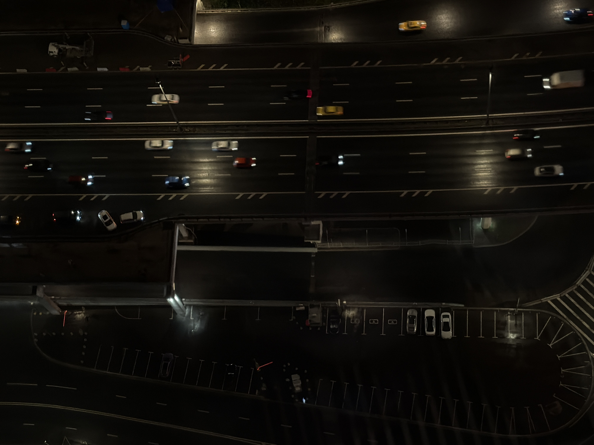 The image captures an aerial view of a highway at night in Khimki, Russia, likely near the Moscow Canal due to the proximity of what appears to be a raised embankment or a bridge structure. The highway features multiple lanes in both directions, illuminated by streetlights and the headlights and taillights of passing vehicles. On the left side of the highway, a dark area with parked cars is visible, suggesting a parking lot or a service area. Several cars are in motion on the highway, their lights creating streaks of color on the wet road surface, indicating recent rain or damp conditions. The scene is characterized by darkness, with artificial lights providing the primary illumination.