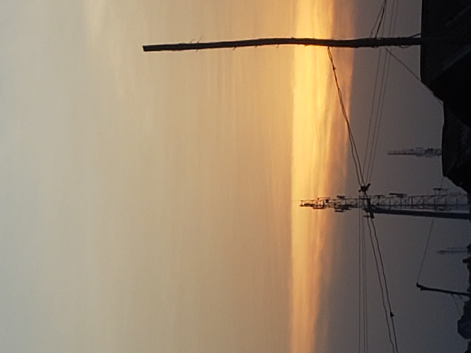The image displays an outdoor scene captured during either sunrise or sunset, dominated by a wide, horizontally banded sky. The sky exhibits a gradient of colors, transitioning from a soft, muted beige or light grey on the left to warm, glowing golden-orange and brownish hues on the right, suggesting the direction of the low sun.
Several tall, dark structures are silhouetted against this colorful backdrop. A prominent, thick utility pole stands vertically on the left side of the frame. Further to the right, a series of metallic lattice towers, likely communication or power transmission towers, are visible, their intricate forms starkly contrasting with the bright sky. Overhead lines, appearing as thin dark lines, stretch across the sky, connecting these structures. The bottom right corner of the image contains a dark, indistinct mass that could be part of a building or another structure.
The overall light is soft and diffused, creating a serene and calm atmosphere. There is no discernible human activity or visible text. The presence of utility poles and towers suggests an urban or semi-urban setting, consistent with the given location context of Adankolo, Nigeria. The scene evokes a sense of tranquil transition, characteristic of early morning or late evening.