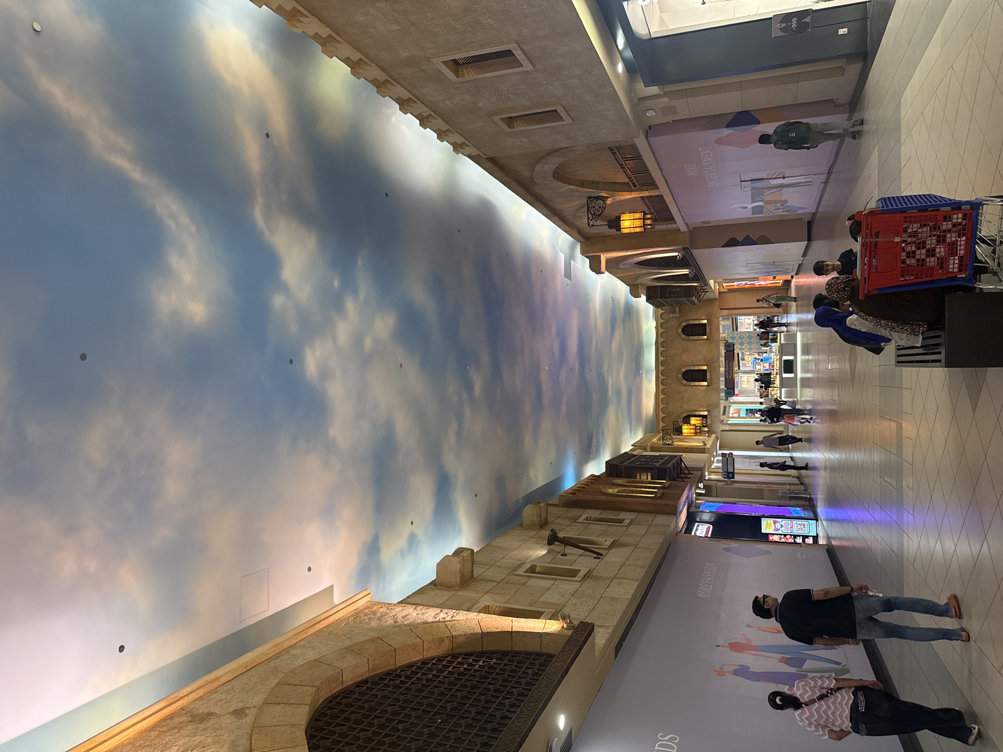 The image shows an indoor marketplace with a high, arched ceiling painted with a sky mural featuring clouds and a sunset. The architecture on either side of the walkway resembles traditional Middle Eastern buildings with stone facades, arched windows, and decorative ironwork. Several people are walking and sitting in the marketplace. In the foreground, a man and woman walk away from the camera. To their right, a group of people sits on a bench with a shopping cart. Further down the walkway, individuals can be seen browsing shops and walking. The lighting is warm, emanating from lamps and the ambient light reflecting off the ceiling. The overall impression is of a vibrant, air-conditioned shopping environment designed to evoke an outdoor bazaar atmosphere. Based on the architectural style, the sky mural, and the overall ambiance, this scene is highly likely to be within the Ibn Battuta Mall in Dubai, United Arab Emirates.