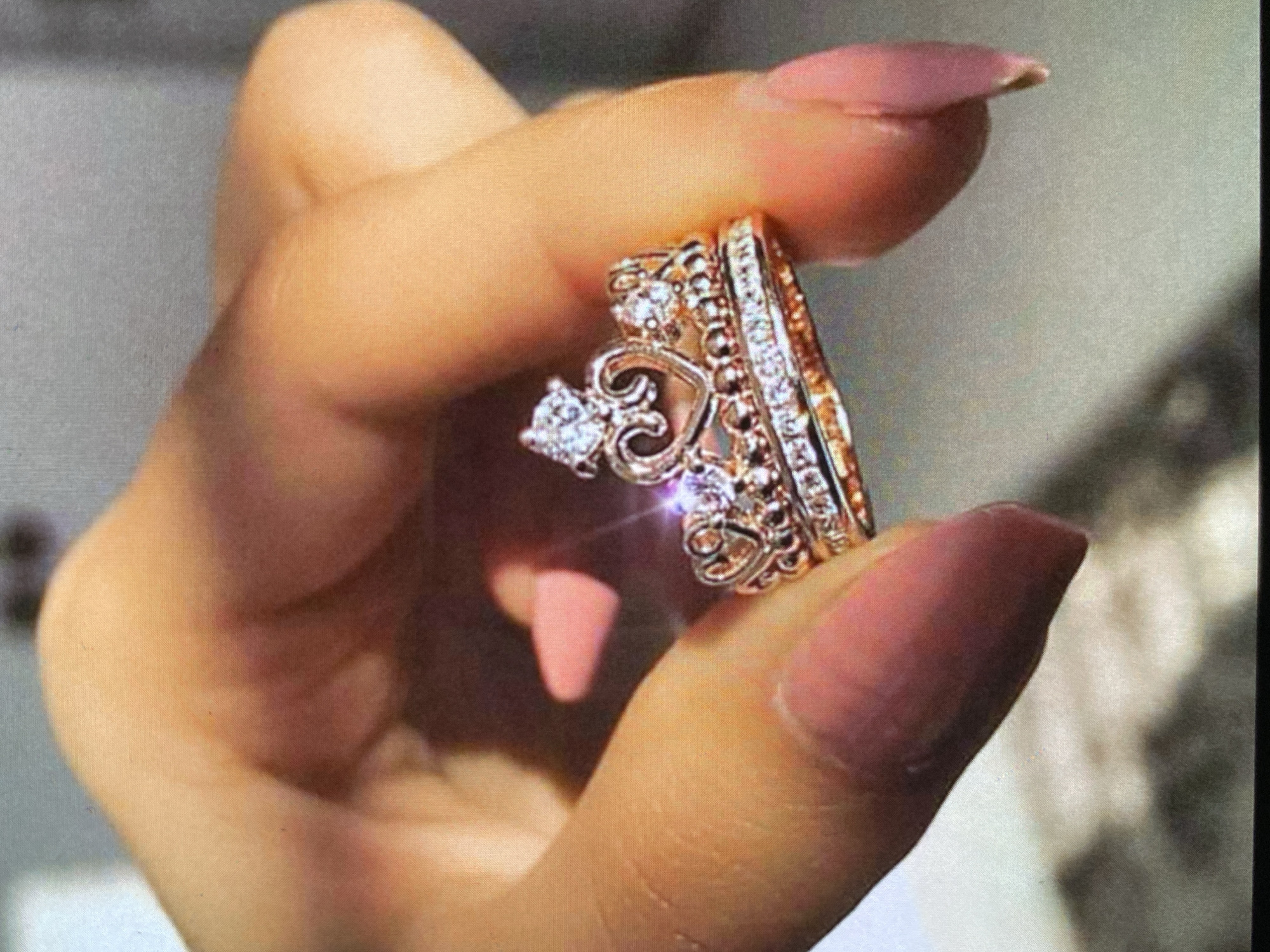A close-up shot features a person's hand delicately holding two ornate rings. The hand, with neatly manicured nails painted in a soft pink or nude shade, positions the rings between the thumb and forefinger for display. Both rings exhibit a warm, gold-toned metal, likely rose gold, and are heavily adorned with sparkling stones.
The more prominent ring has a distinctive design, featuring a heart-shaped setting that resembles a small crown or tiara. It showcases a larger, brilliant central stone flanked by multiple smaller stones, all contributing to its intricate, jeweled appearance. The second ring is a slender band, appearing to be an eternity or half-eternity style, completely encrusted with small, glittering stones that catch the light.
Bright, direct lighting illuminates the jewelry, causing numerous sparkling reflections on the stones, highlighting their brilliance. The background is softly blurred into a light, neutral tone, suggesting an indoor setting and emphasizing the rings as the primary subject. The image's purpose is to showcase the intricate details and radiant sparkle of the jewelry. There is no visible text or overt emotional expression. This scene is set in Bankya, Bulgaria.