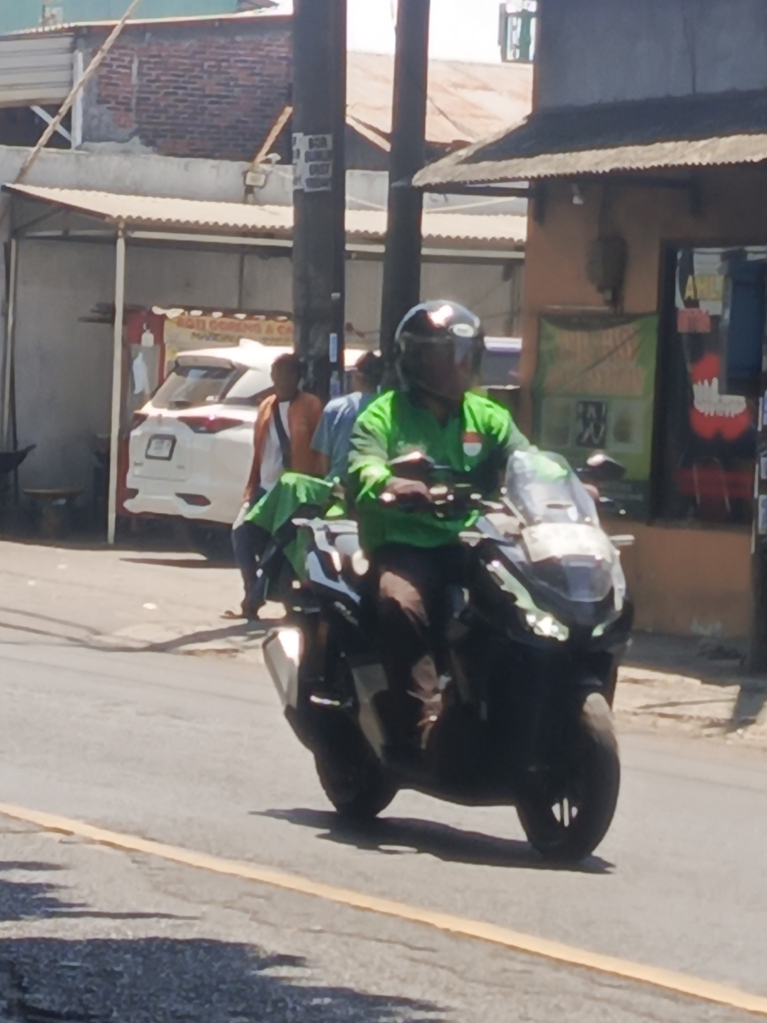 A man wearing a green shirt and a black helmet rides a black motorcycle with a large windshield. He is wearing dark pants and appears to be on a road in an urban or semi-urban area. To the left of the motorcycle, a white car is parked in front of a building with a corrugated metal roof. Behind the car, a person in an orange jacket is visible. The background includes buildings with various roofing materials and a utility pole. The scene is illuminated by bright sunlight, casting shadows on the road. The image appears to have been taken in Kedungrejo, Indonesia.