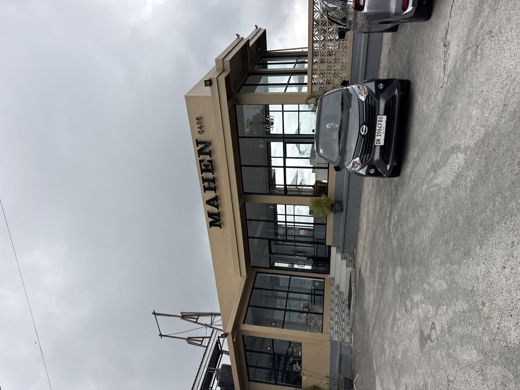 The image depicts the MAHEN CAFE, a contemporary structure located in Kintamani, Indonesia. The cafe features a light tan facade with extensive glass windows, offering a view of the interior, which includes seating areas and a chandelier. The name "MAHEN CAFE" is prominently displayed in black lettering on the front of the building. In the foreground, two cars are parked on a paved area. The car closer to the viewer is a dark gray hatchback with the license plate "DK 1964 FBO". Behind it, partially visible, is a silver SUV. The ground is a rough, gray asphalt. A decorative patterned concrete wall runs along the side of the building. The sky is overcast with a light gray, cloudy appearance, and a thin black wire stretches across the upper portion of the frame, with a small pink object possibly attached to it. A bamboo structure, possibly part of a kite flying setup, is visible to the left of the cafe, against the cloudy sky. The overall environment suggests an outdoor setting in a town.