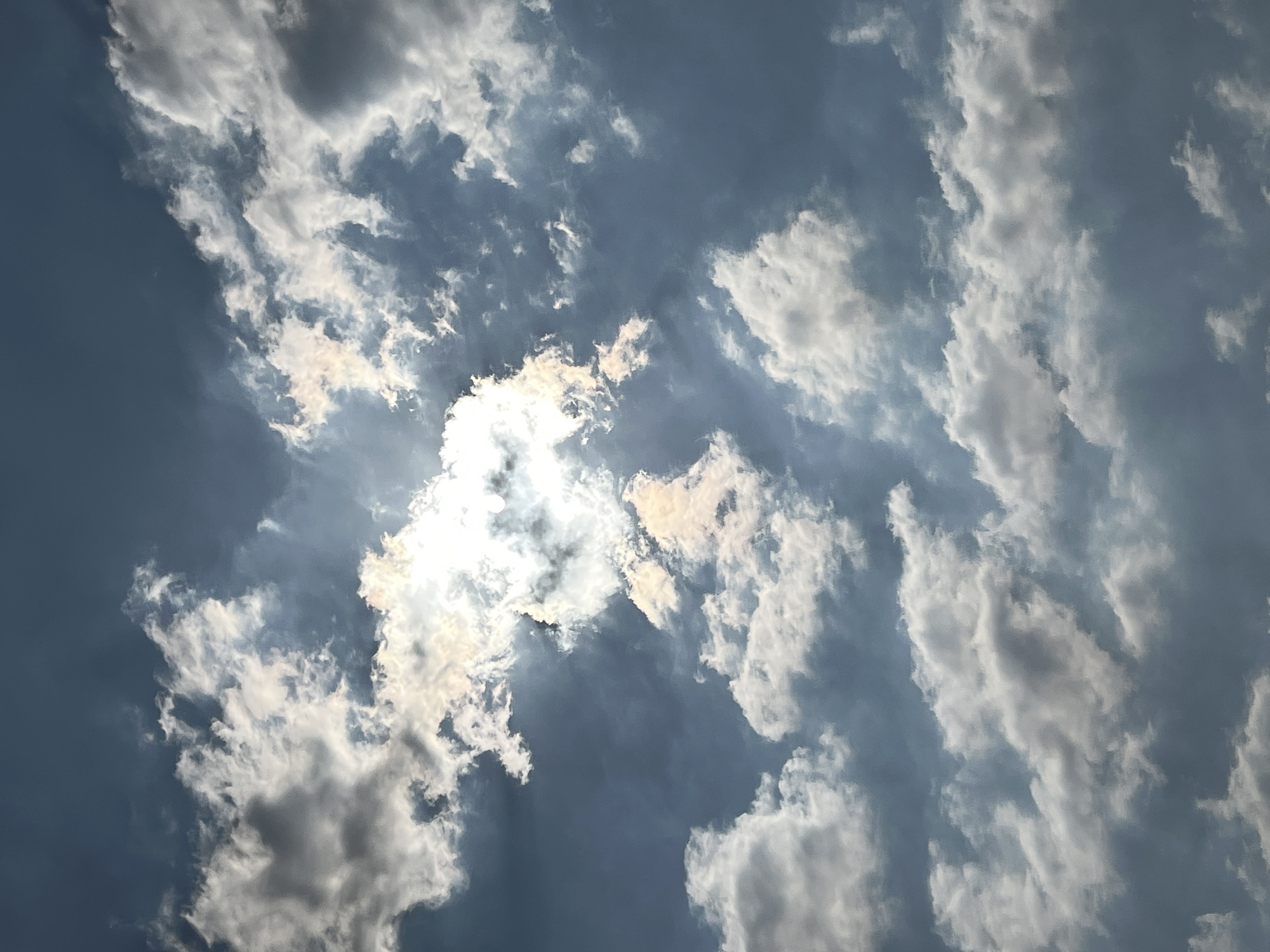 The image is a close-up, low-angle shot of a partly cloudy sky. The sun is partially obscured by clouds, creating a bright, glowing halo effect. The clouds are a mix of bright white and darker gray, with some showing hints of orange and yellow from the sunlight. The sky is a deep blue, indicating it is daytime. The scene appears to be an outdoor environment. There are no visible people, objects, or actions taking place. The image evokes a sense of atmospheric beauty and dynamic weather conditions. There is no information within the image or provided context to ascertain the specific location in Bandung City, Indonesia.