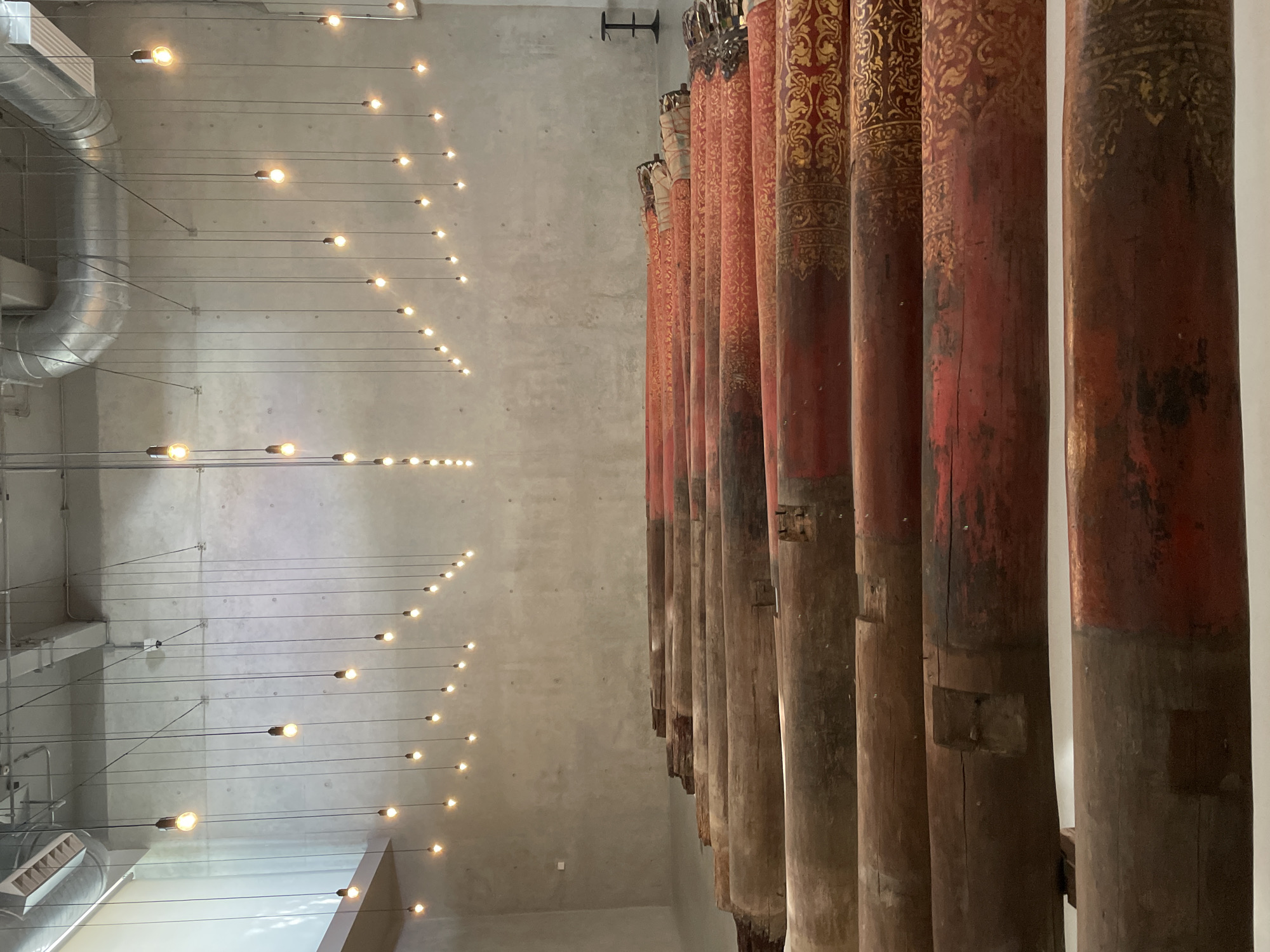 The image depicts an interior space in Bangkok, Thailand. A row of ornately decorated, cylindrical wooden columns stands against a gray concrete wall. The columns feature reddish-brown paint with gold embellishments near their tops. To the left of the columns, a pattern of evenly spaced pendant lights hangs from the ceiling, their thin wires stretching across the gray concrete surface. Metal ductwork is visible among the lights near the ceiling. The overall lighting is soft and diffused.