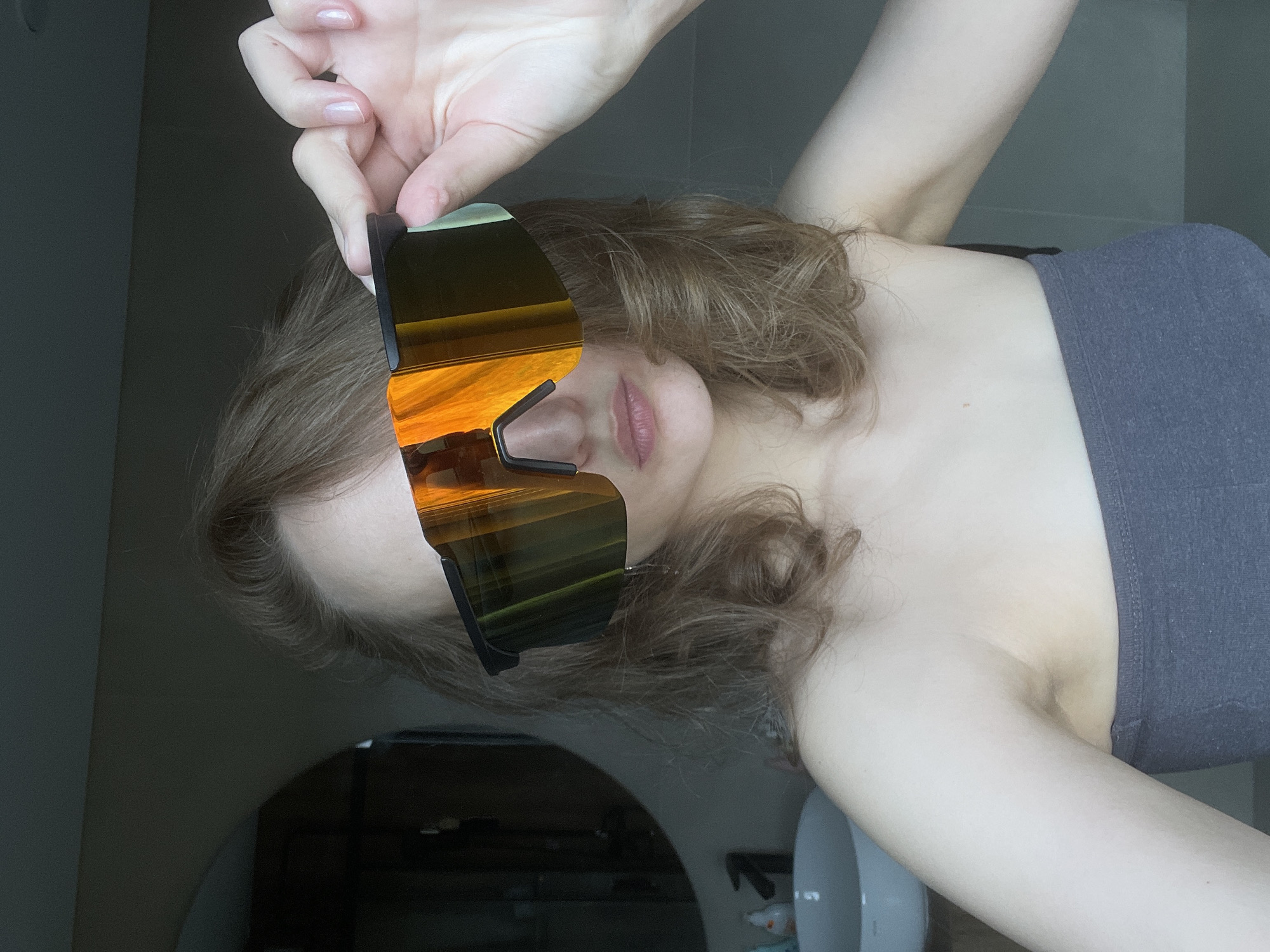 A woman with light skin and curly brown hair is holding up large, reflective sunglasses in front of her face. The reflection in the sunglasses shows an orange sky, possibly at sunset or sunrise. The woman is wearing a gray top. The background is a room with a mirror reflecting an unknown space. A white sink and various bottles are visible in the periphery. The location is inferred to be Подшивалово, Russia.