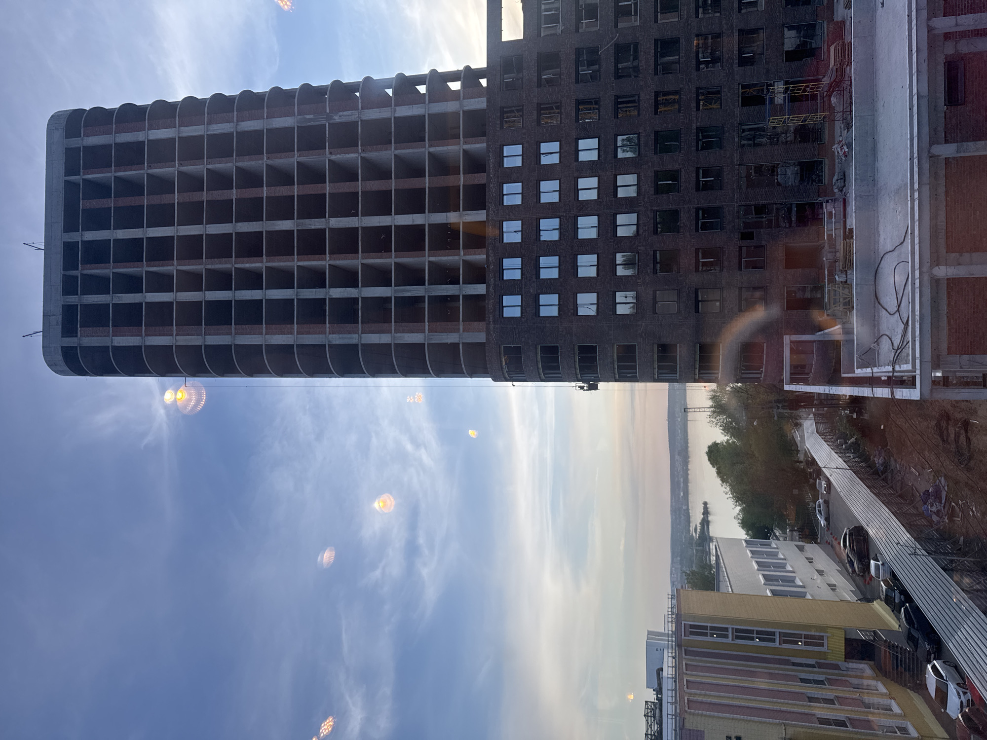 The image depicts a view from a window in Izhevsk, Russia, showcasing two buildings. The building on the left is an unfinished high-rise with a grid-like structure of empty window frames. It has a rounded edge and is constructed of brick and concrete. To the right, there is a completed building with many windows, darker brick, and some scaffolding present. Below these two buildings is a vista of a body of water, possibly a river, with the skyline visible in the distance. Several other buildings of various sizes and colors, along with some trees, are visible closer to the foreground. There is a street with parked cars along the lower edge of the image. The sky is light blue with some scattered clouds. The image appears to be taken during daytime with ample lighting. There is some sun glare/flare visible in the sky.