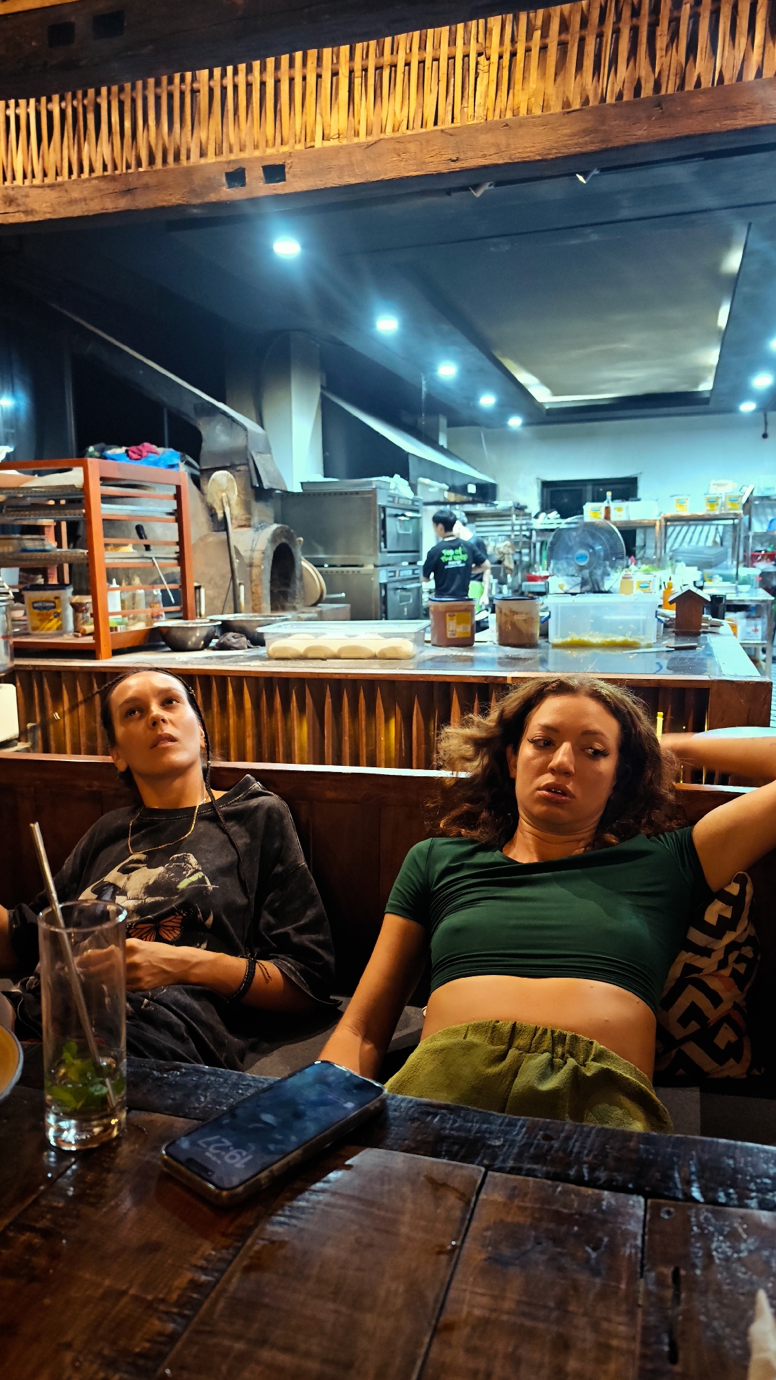 Two women are seated at a wooden table in a restaurant in 1 Ward, Hà Giang, Vietnam. The restaurant features an open kitchen visible in the background, including a pizza oven and stainless steel cooking surfaces. One woman, on the left, wears a dark hoodie and sits beside an empty glass with a straw. The second woman, on the right, wears a green top and olive-colored pants. A smartphone is placed on the table between them. The restaurant has warm, ambient lighting and a decorative wooden structure on the ceiling.