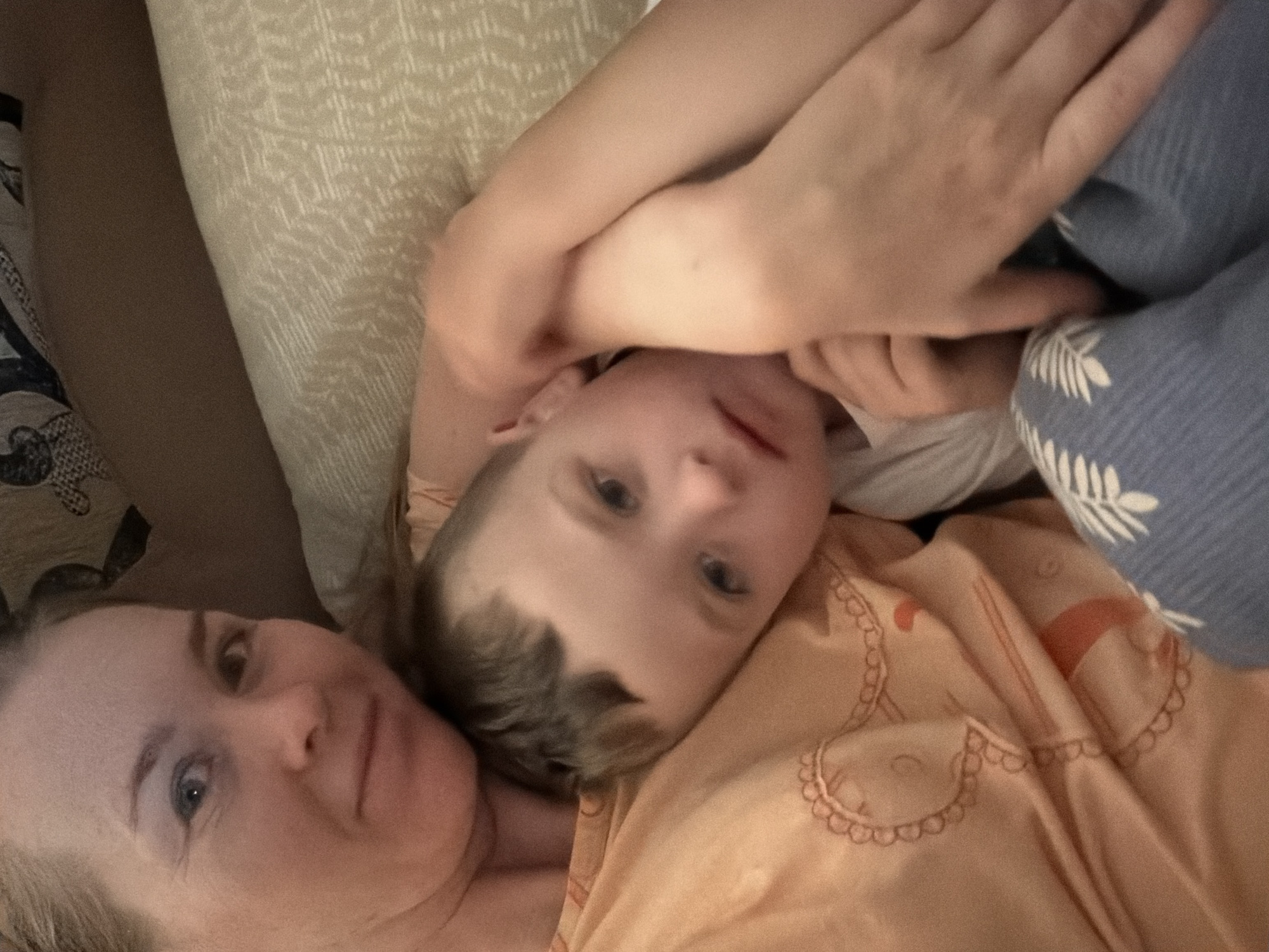 This is a close-up, overhead shot of a mother and child lying down together. The mother's face is on the left side of the frame, with her eyes open and looking towards the camera. Her hair is blonde and appears to be pulled back. The child is positioned above her, with their head resting on her chest and arm. The child has light brown hair and blue eyes and is looking directly at the camera with a calm expression. They are both wearing casual clothing. The child is wearing a light-colored t-shirt, and the mother is wearing an orange shirt with a decorative pattern. They are lying on a bed with light-colored bedding and a patterned blanket. The background suggests an indoor setting, possibly a bedroom, with soft lighting. The overall mood is one of intimacy and comfort.