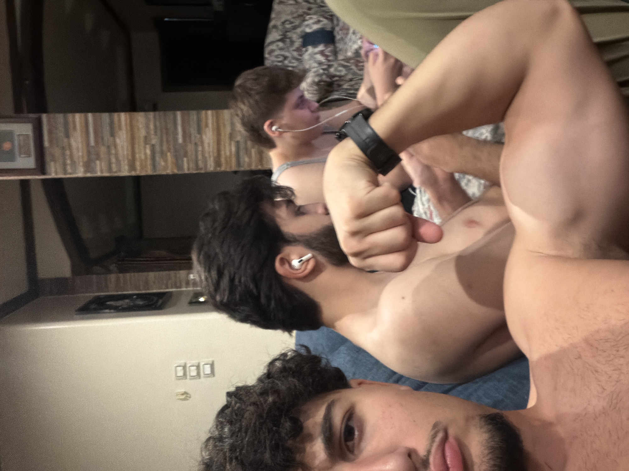 The image depicts three young men in what appears to be a living room. The man in the foreground, positioned on the left, is partially visible, showing his face, shoulder, and bicep. He has curly dark hair, a goatee, and is wearing an airpod in his right ear. He is flexing his bicep, and his arm is adorned with a black watch. Two other men are visible behind him. The man in the middle has dark hair and is also wearing an airpod. The man on the right has lighter brown hair and is wearing earbuds, looking down at a device. The room's background suggests an indoor setting with a couch, a patterned sofa, and walls with decorative stonework and framed pictures. Electrical outlets are visible on the wall. The lighting is dim, suggesting an evening or indoor setting. The location is identified as being in the city of Sarouja Municipality, Syria.