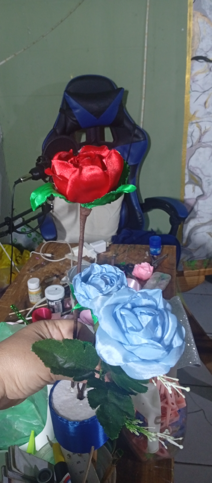 A hand holds a floral arrangement consisting of two light blue and one red silk rose with green leaves in front of a cluttered workbench and a black and blue gaming chair. The background includes craft supplies such as bottles of paint, ribbon, and other materials, against a light green wall with a painted design. The location of Juan Griego, Venezuela, cannot be determined from the visual information.