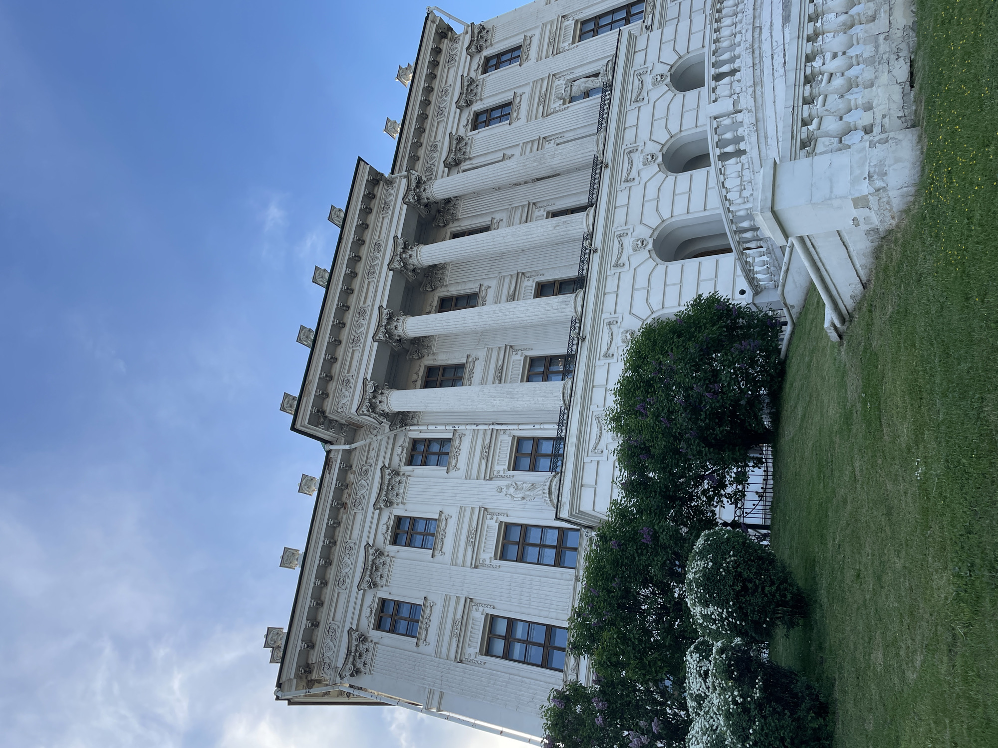 The image shows a white building with columns in Moscow, Russia. The building has a complex architectural design with decorative elements and multiple windows. In front of the building is a green lawn and bushes. The sky is blue with some clouds.