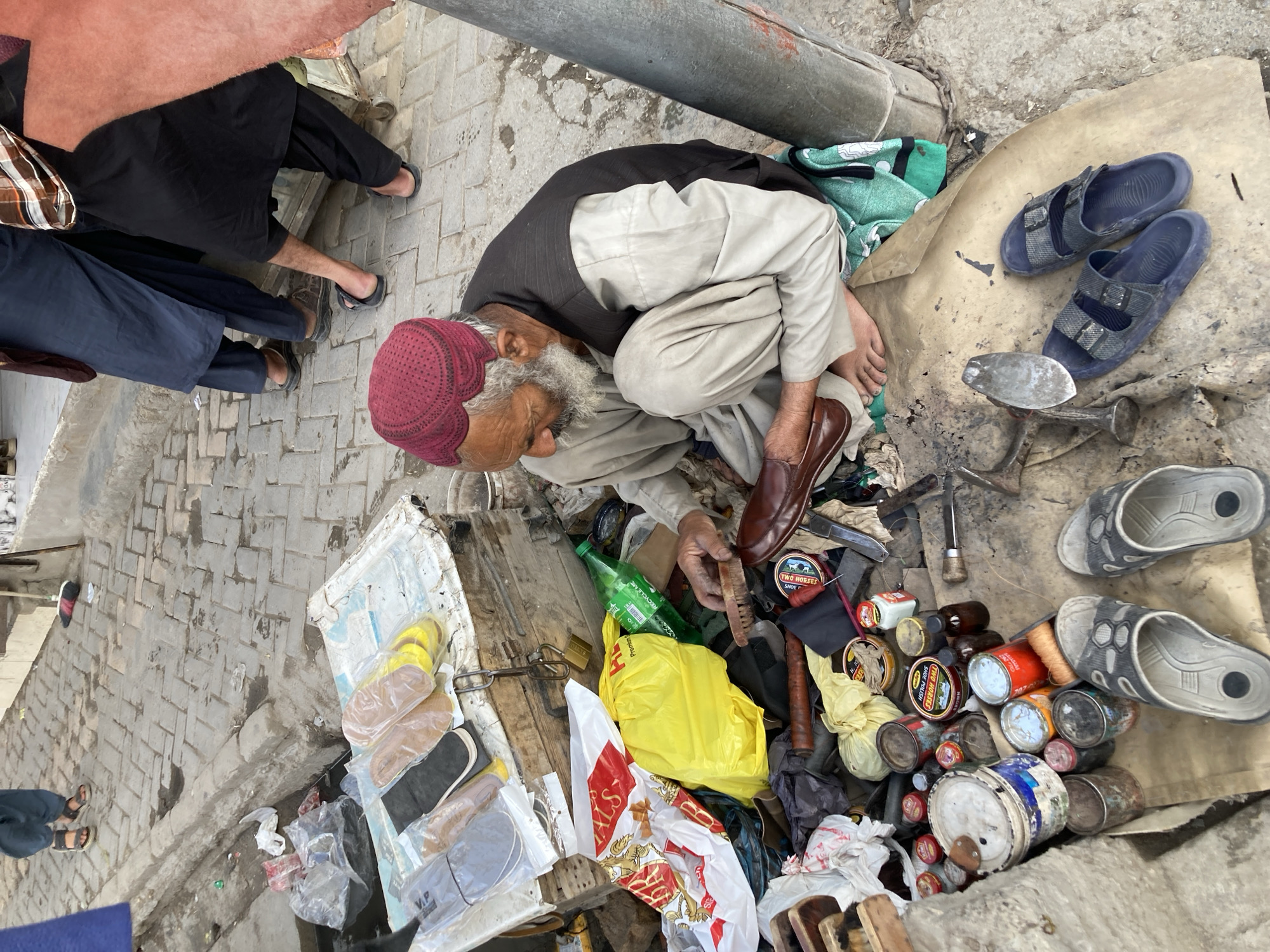 The scene depicts an elderly shoemaker in Quetta City Tehsil, Pakistan, focused on polishing a brown shoe. He wears a red cap, light-colored clothing, and sits cross-legged amidst his tools and supplies. Paint cans, other shoes, and various objects litter the ground around him on a makeshift workspace covered with cloth. People walk by in the background. The environment appears to be an outdoor setting with stone-paved surfaces.