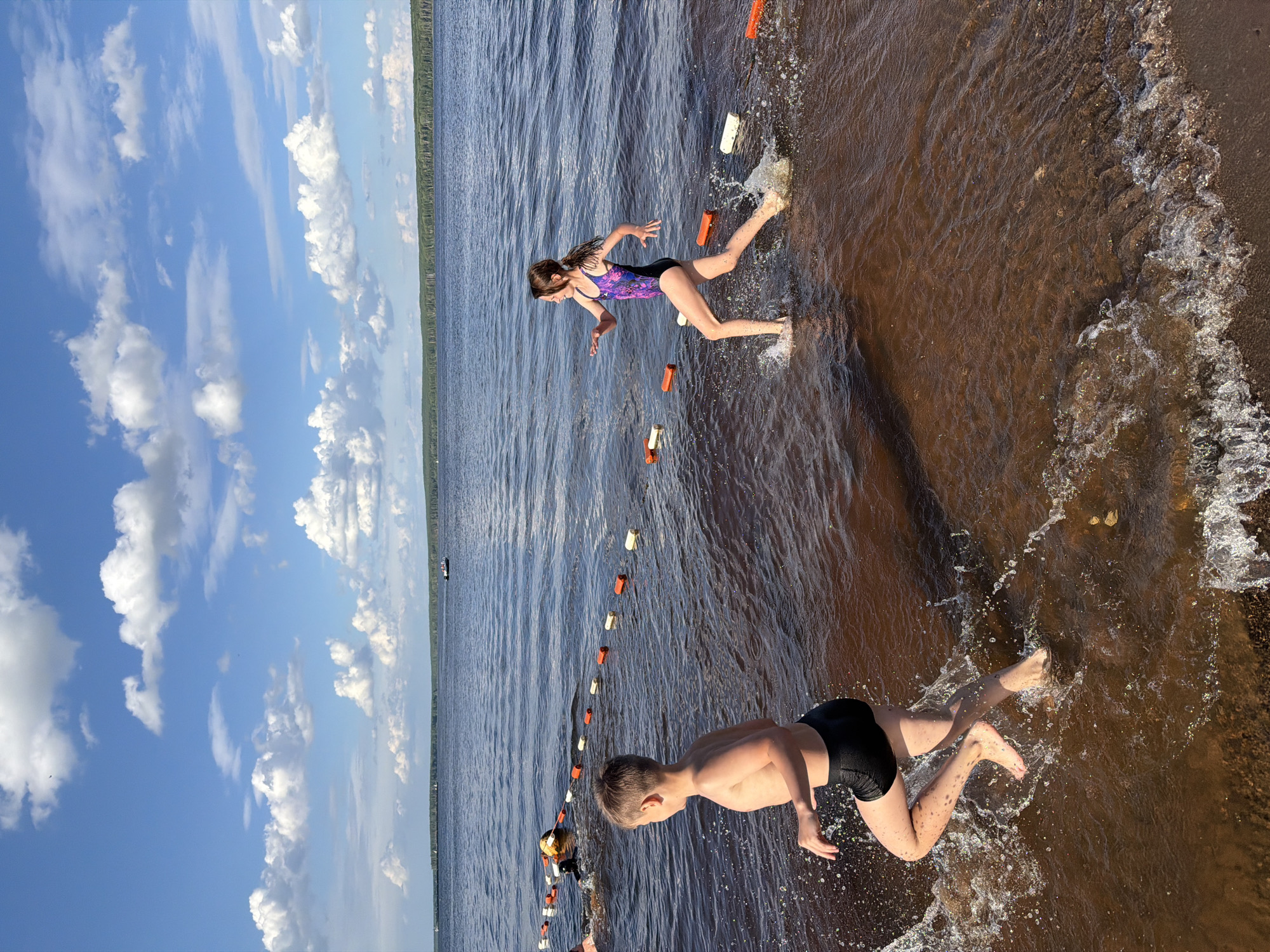 This media file shows two children playing in the water at a beach. One child, a girl in a purple and black swimsuit, is in the water, appearing to be running or jumping. The other child, a boy in black swim trunks, is also in the water, splashing with his hands. They are near a rope with orange and white buoys in the shallow water. The scene is set on a sandy beach with brown water. In the background, there is a line of trees and a body of water, possibly a lake or river, with a boat in the distance. The sky is blue with scattered white clouds. The image is taken from a slightly elevated angle, with the horizon visible in the upper portion of the frame. The time of day appears to be daytime, with bright sunlight casting shadows. The location context is Chaikovsky, Russia.