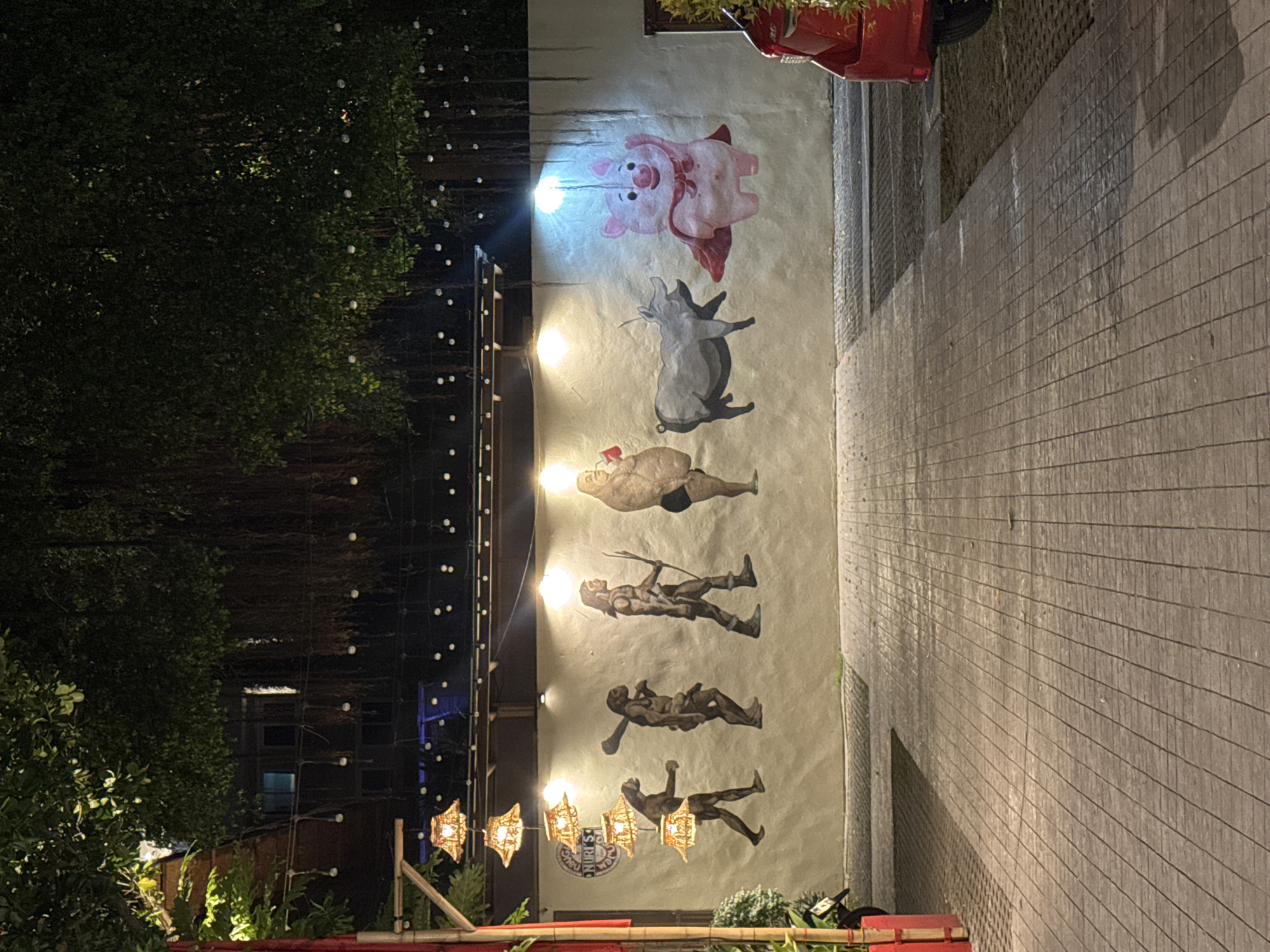 The image depicts a mural on a white wall in Patong, Thailand, likely on a street or walkway. The mural displays a progression of figures, starting from what appears to be early hominids, progressing through various stages of evolution that end with a cartoonish pig wearing a red cape and mask, waving. The figures are painted in earthy tones. Above the mural, there are string lights and lush greenery hanging from above, suggesting an outdoor or semi-outdoor setting. In the foreground, a paved walkway is visible, and to the right, the rear of a red car is parked. The lighting suggests it is evening or nighttime. A sign with the text "NURI'S" is partially visible on the wall to the left of the mural.
