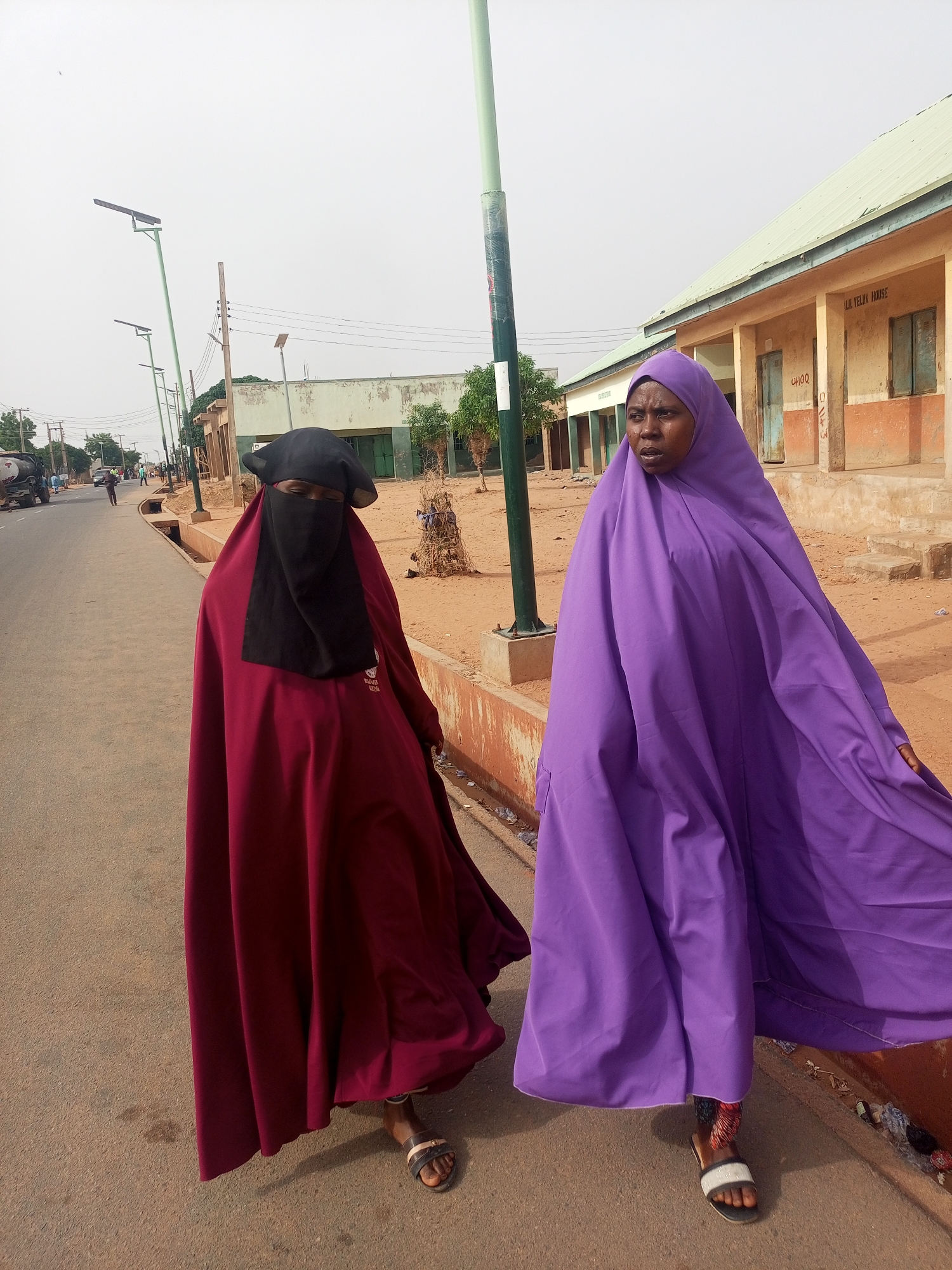 Two women wearing flowing garments walk along a paved street in Gombe, Nigeria. The woman on the left wears a dark red garment with a black face covering. The woman on the right wears a purple garment. Both women wear sandals. A building with a light green roof and the words "I.H. YELWA HOUSE" is visible in the background. Streetlights line the road. Trash is scattered along the curb. Other buildings and trees are visible further down the street.