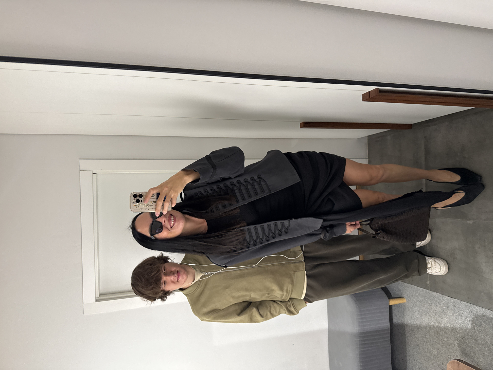 This image is a mirror selfie taken in what appears to be a fitting room, possibly a boutique or a department store. A woman with long dark hair, wearing sunglasses, a dark jacket with decorative braiding, and a black dress, is holding a smartphone to take the picture. She is smiling. To her left stands a young man with brown curly hair, wearing a light olive green bomber jacket over a cream t-shirt and dark pants. He has white earbuds in. They are both looking towards the camera. The background shows a white wall, a doorway, and a portion of a full-length mirror. A grey bench with wooden legs is visible to the left of the frame. The floor is a dark grey tile. There is no visible indication of the location being in Chaikovsky, Russia.