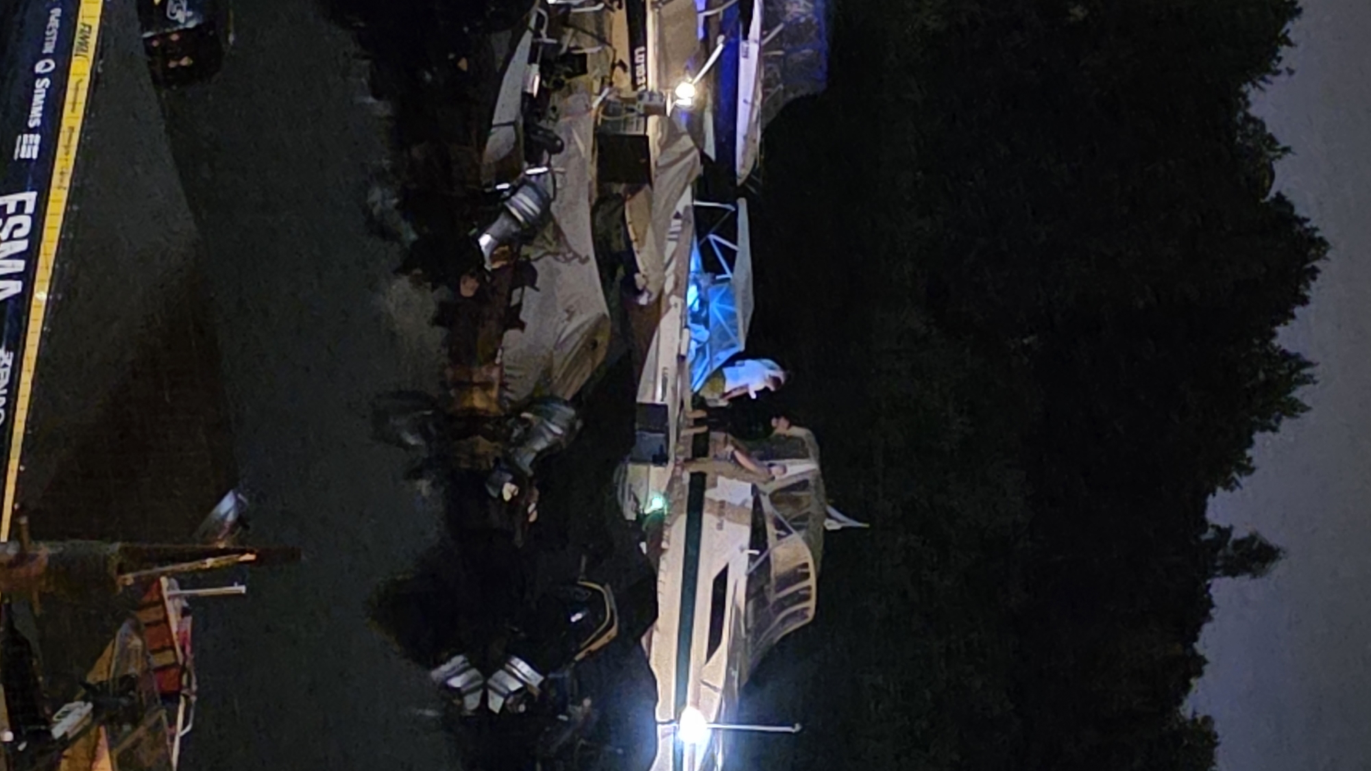 At night, a group of people are standing on or near several boats docked on a body of water. The boats are of various sizes and styles, with some covered and others open. One boat has a blue tarp over part of it. The background consists of a dark treeline against a night sky. The scene appears to be a marina or harbor. The image does not provide specific details to identify the location as Riga, Latvia.