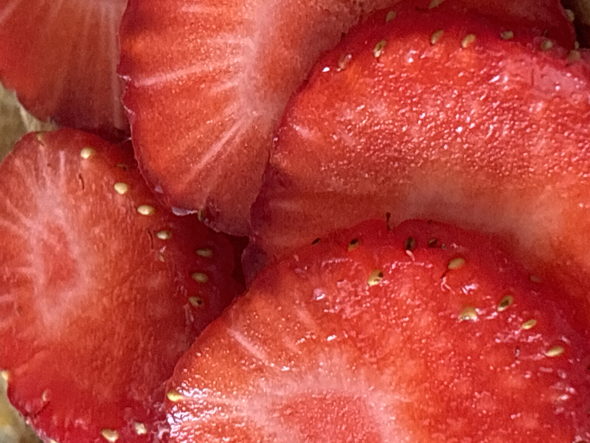 The image is a close-up, overhead view of several sliced strawberries. The strawberries are bright red with visible seeds scattered throughout their flesh. The lighting highlights the juicy texture of the fruit, creating a vibrant and detailed image. The background appears to be a light brown, textured surface, possibly a cutting board or a similar food preparation surface. The image does not contain any elements that would suggest a specific location in Sochi, Russia.
