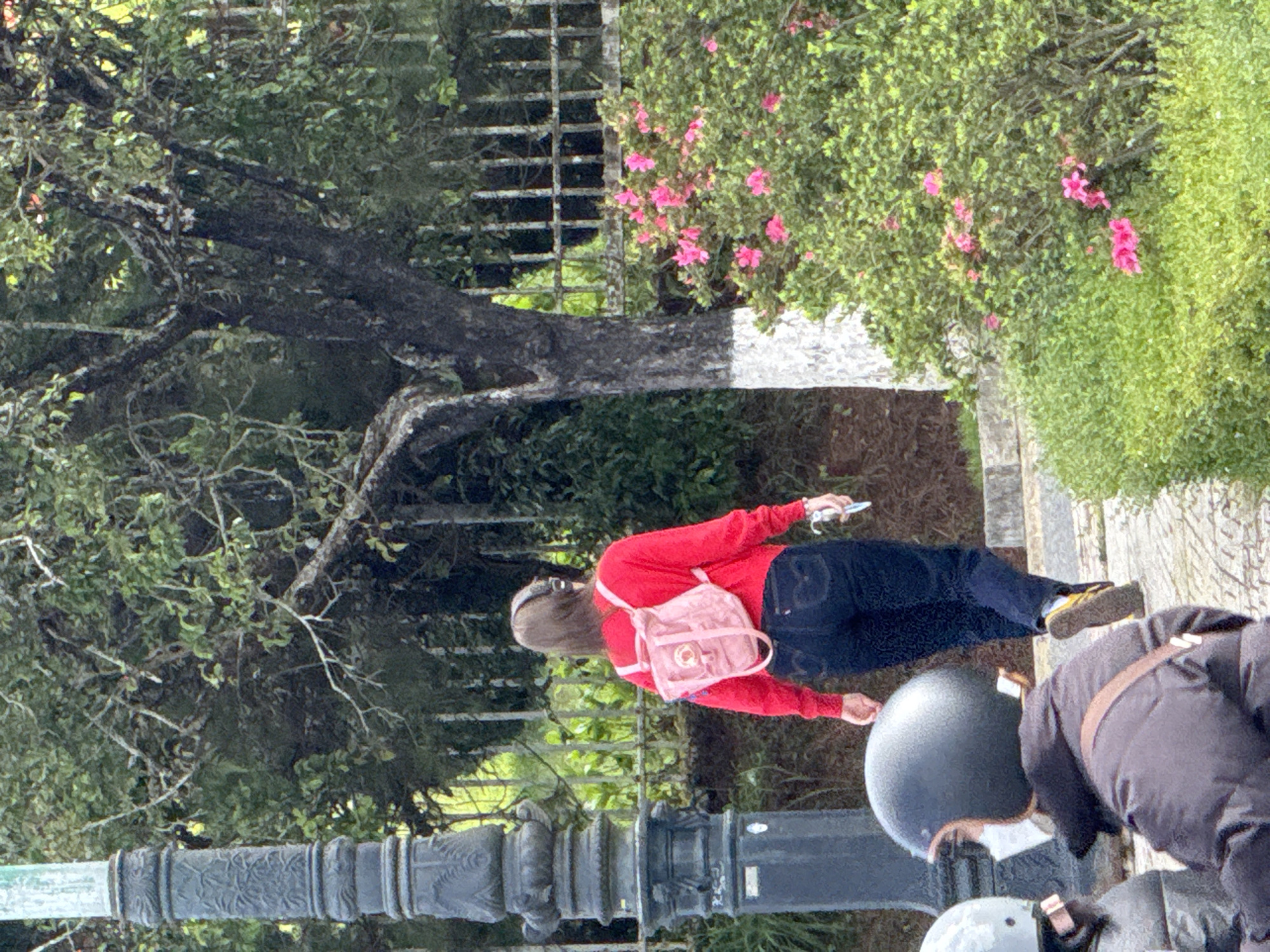 A woman walks away from the camera on a paved walkway in Dalat, Vietnam. She wears a bright red sweater, dark blue jeans, and a pink backpack. Headphones are visible around her neck. In the foreground, two people on a scooter, one wearing a helmet, are visible from behind. The environment includes a decorative lamppost, a metal fence, trees, and flowering bushes with pink blossoms. The ground is paved with stones.
