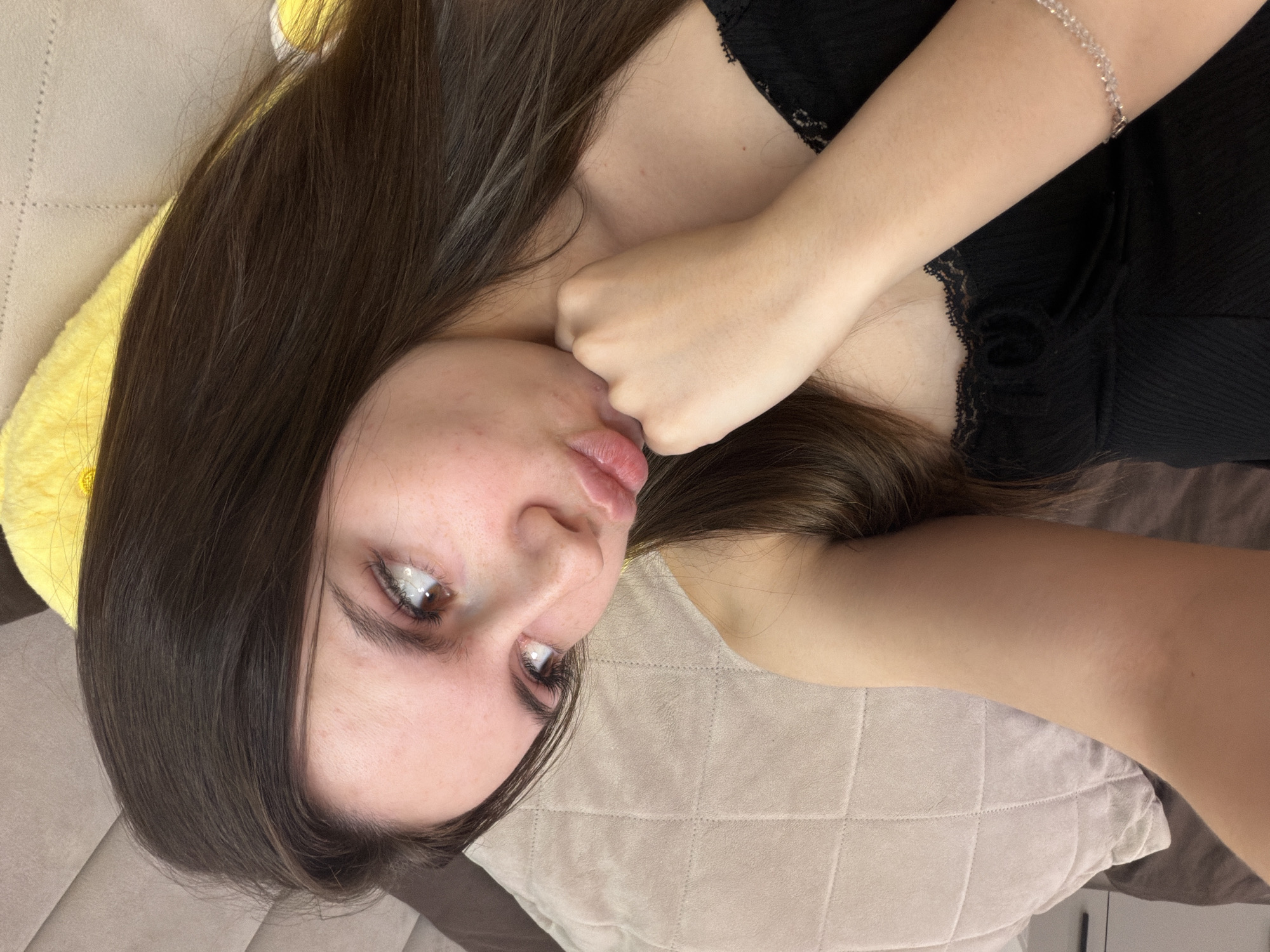 A young female with long, dark brown hair is depicted lying on an upholstered surface, possibly a sofa or bed, in an indoor setting. She is looking upwards and slightly to her left, with her lips pursed into a puckered expression. Her right hand is balled into a fist and positioned under her chin/jawline. Her left arm extends towards the upper left of the frame, adorned with a light-colored beaded bracelet on her wrist. She is wearing a black, ribbed top featuring lace trim around the neckline. The background includes a light beige quilted pillow behind her head, a darker brown pillow partially visible in the upper left, and a bright yellow soft object to her right. The image provides no visual indicators to determine its specific geographical location, such as the city of Chaikovsky, Russia.