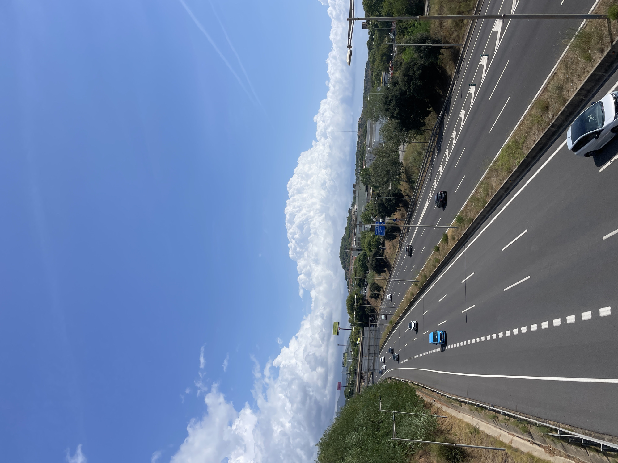 A high-angle medium shot captures a multi-lane highway on a sunny day with a bright blue sky and scattered white clouds in Badalona, Spain. The highway stretches into the distance, with traffic flowing in both directions. Several cars are visible on the road, including a blue van in the foreground. Trees and vegetation line the sides of the highway, and buildings and hills can be seen in the background. Road signs and lampposts are also present. The scene appears to be a typical urban or suburban highway environment.