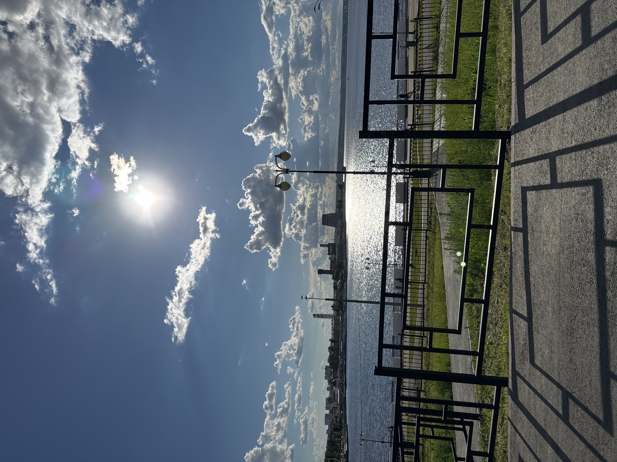 The image shows a view of the Izhevsk Pond and embankment in Izhevsk, Russia, on a sunny day with a clear blue sky and scattered white clouds. The sun is shining brightly, creating a glare on the water's surface. In the foreground, there's a black metal fence casting shadows on the paved embankment. Behind the fence, there is a strip of green grass. In the middle ground, the calm water of the Izhevsk Pond reflects the sunlight, with the city skyline visible in the distance. The cityscape includes buildings of various heights and architectural styles, as well as a few industrial structures like smokestacks and towers. Street lights are positioned along the embankment. There are no visible people in the image.