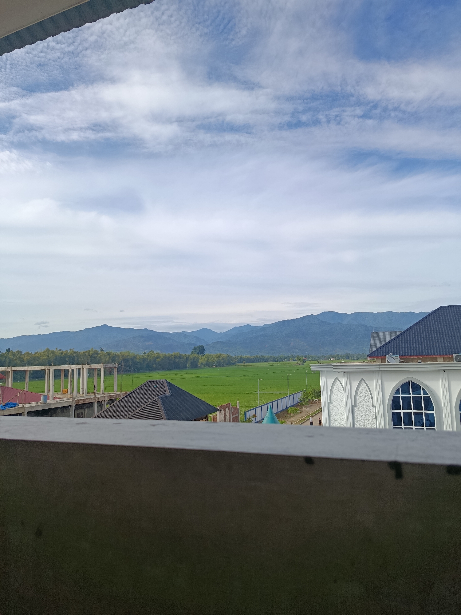 The image depicts a rural landscape with buildings and mountains in the background. In the foreground, a gray railing is visible. To the left, an unfinished concrete building with pillars stands under a blue sky with scattered clouds. In the middle ground, lush green rice paddies stretch towards a line of trees, backed by a range of blue-hued mountains. To the right, a white building with arched windows and intricate patterned decorations is partially visible, suggesting it could be a mosque. The overall scene appears to be an exterior view, possibly from an elevated position.