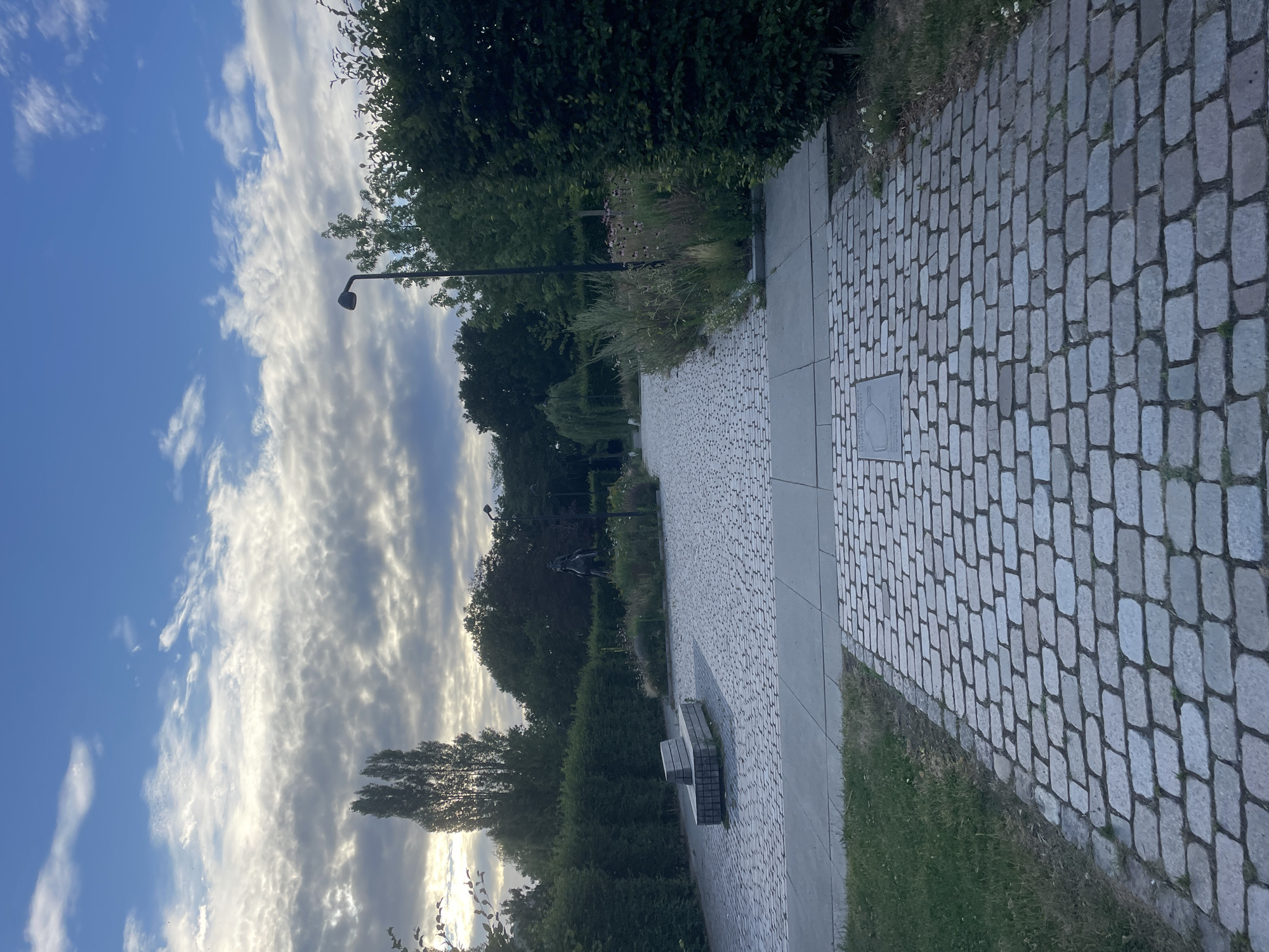 The image displays a park pathway made of cobblestones, curving into the distance. To the right, the path is bordered by a strip of green grass. Trees and lush greenery line the left side of the pathway, with a street lamp visible amidst the foliage. In the background, a statue or sculpture can be seen within the park. The sky above is a mix of blue and dramatic, streaked clouds, suggesting either early morning or late afternoon. The overall mood is peaceful and serene, evoking a sense of quiet contemplation in a natural setting. There is no visible text in the image.