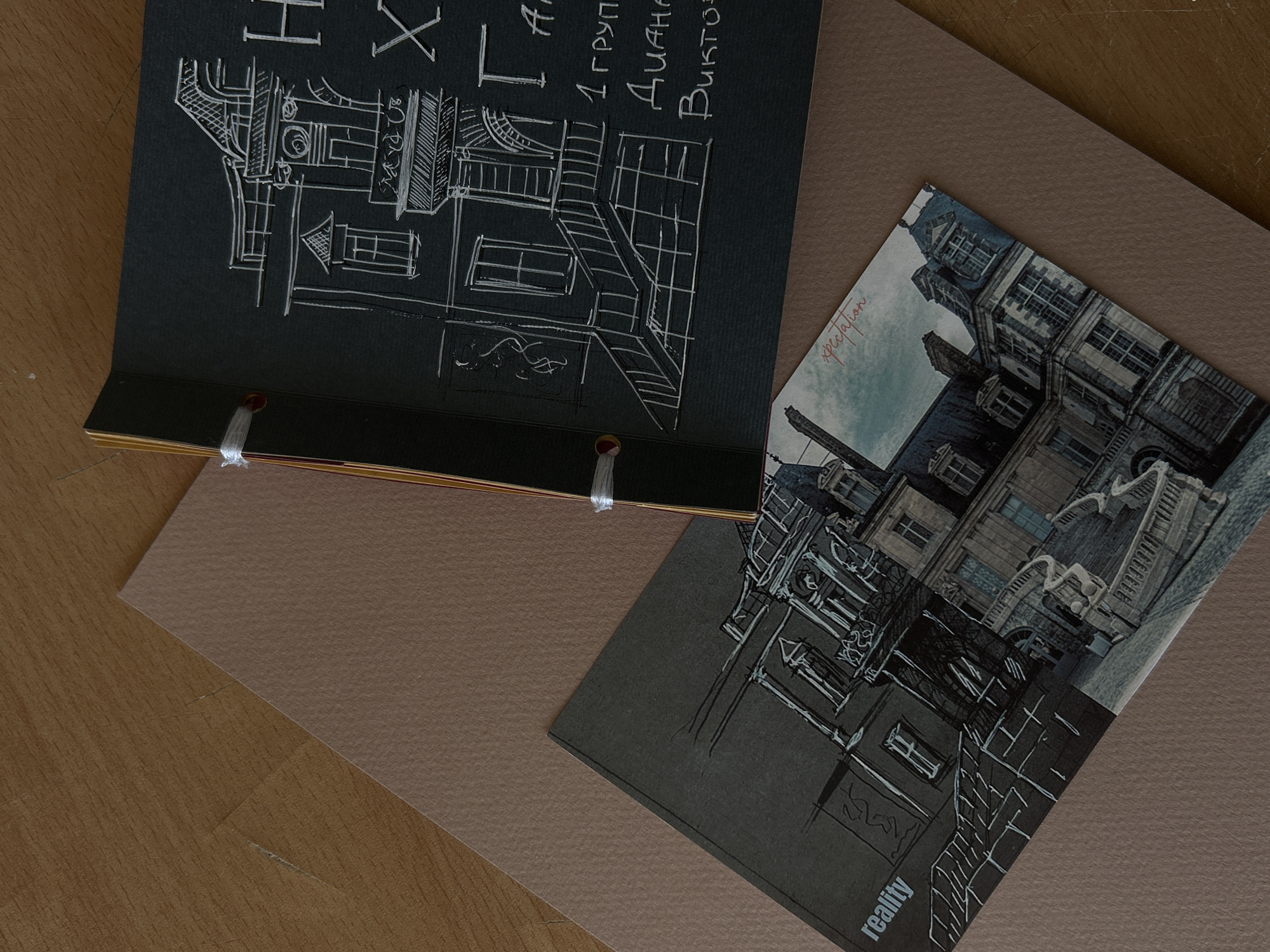 The image shows two black sketchbooks and a light brown piece of paper laid out on a wooden surface. The top sketchbook is open, revealing a white chalk-like drawing of a building facade with an ornate staircase and windows. Text is written in white in the right portion of the page. The bottom sketchbook is closed and shows a similar architectural drawing. A postcard with an image of a chateau and the word "expectation" written in red is placed on top of the bottom sketchbook, partially overlapping it. The word "reality" is visible in white text on the lower left corner of the bottom sketchbook. The scene is lit from above, casting shadows. There are no people in the image. The context does not provide any information about the location in Sofia, Bulgaria.