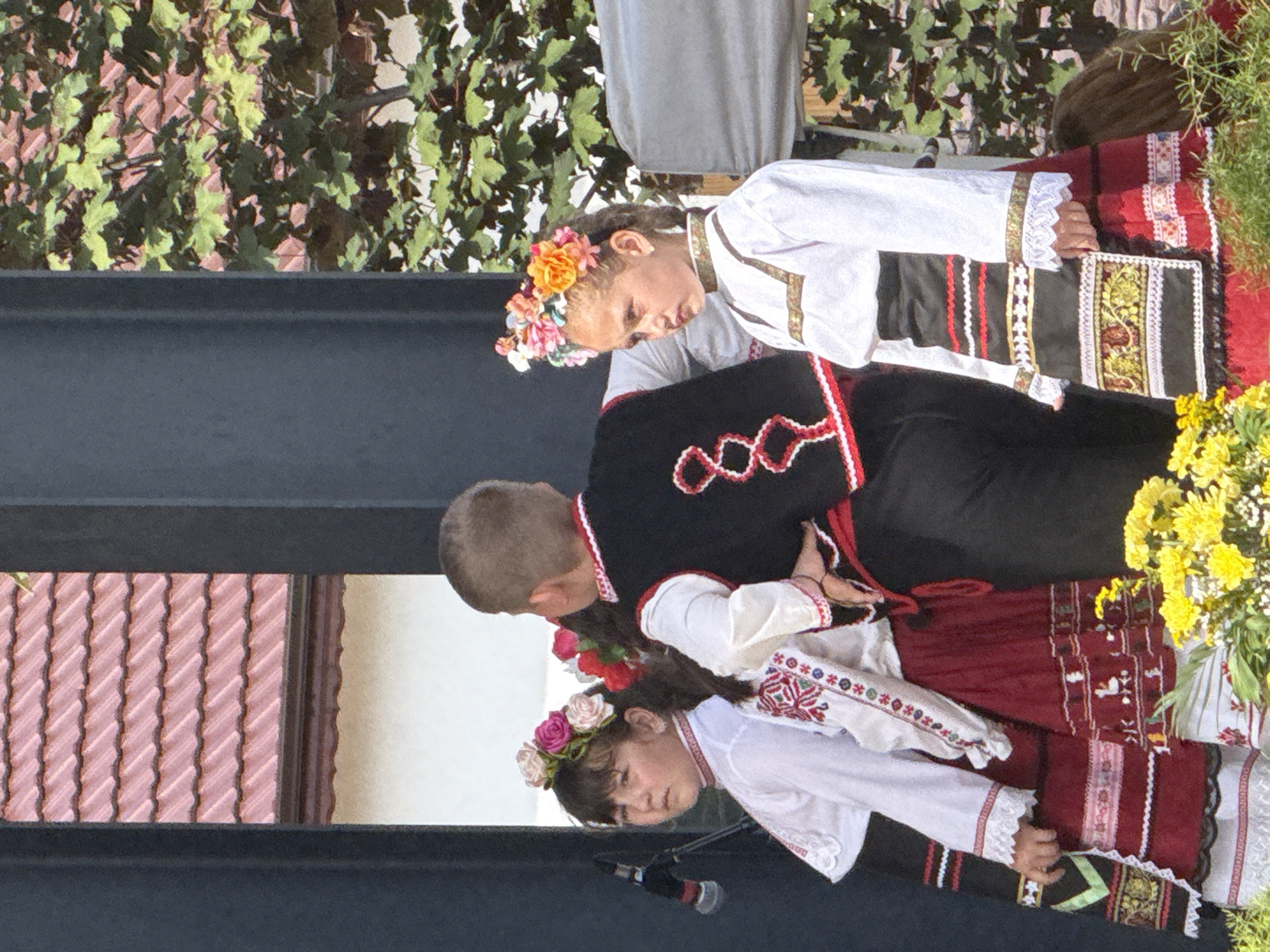 A group of children are dressed in traditional Bulgarian folk costumes on an outdoor stage. The children, two girls and a boy, are positioned from left to right, with the boy in the center. The girl on the left has dark hair styled with a floral wreath of pink and white flowers. She wears a white embroidered blouse with a red skirt and apron. The boy in the middle has short brown hair and wears a black vest with red and white trim over a white shirt, and dark trousers. The girl on the right has braided dark hair adorned with a colorful floral wreath. She is dressed in a white embroidered blouse and a red skirt with a patterned apron. They appear to be participating in a performance, possibly a folk dance or cultural event. In the background, a microphone is visible to the left, and a tiled roof and foliage can be seen. The image was taken in Pordim, Bulgaria, at an event on the Town Square.