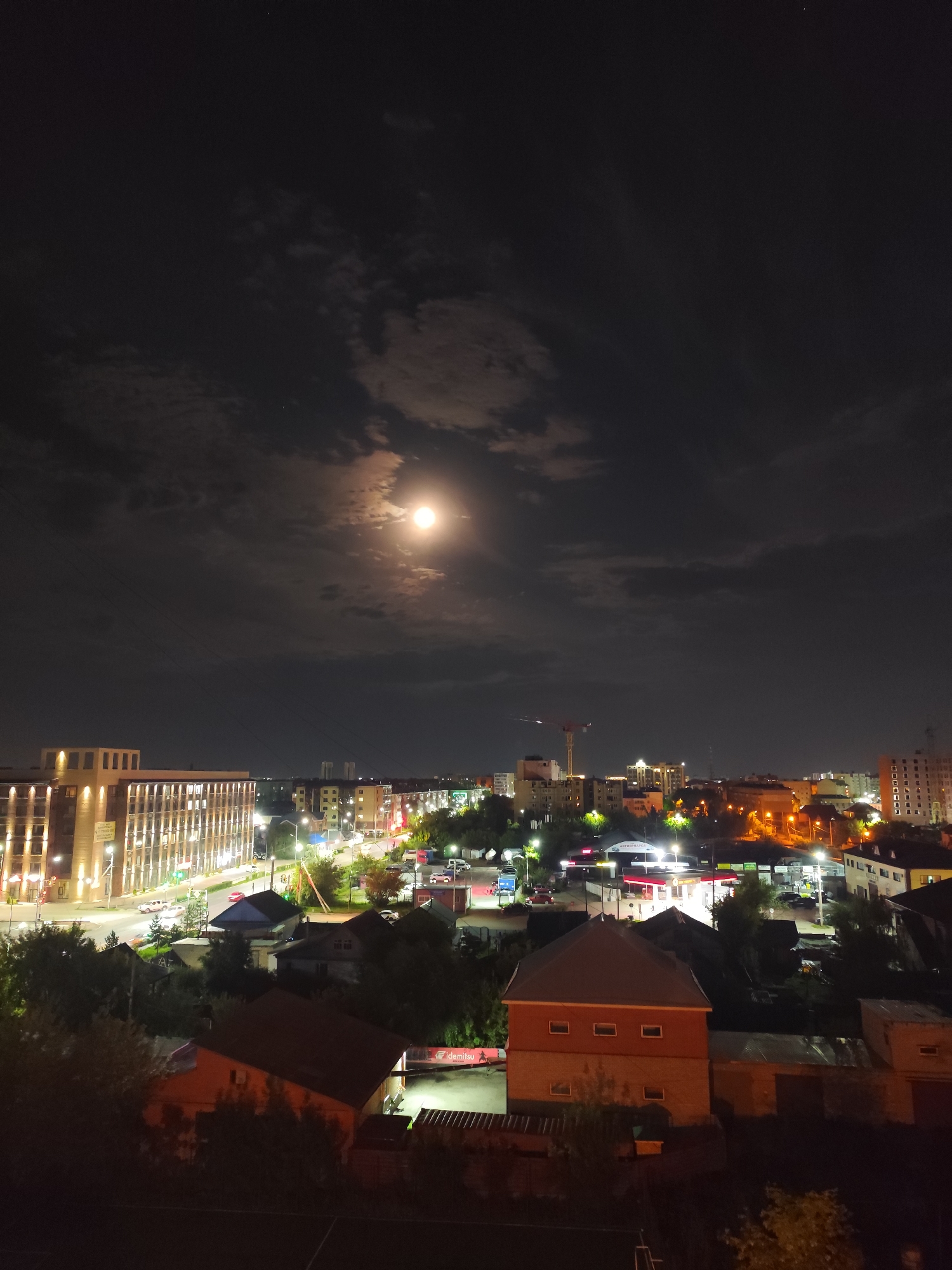 This is a nighttime aerial photograph of a city, likely Petropavl, Kazakhstan, illuminated by a full moon and artificial lights. In the foreground, several residential buildings with red roofs are visible, some with visible windows and doorways. Beyond these, a street scene unfolds with cars, streetlights, and commercial buildings, including one large building with numerous illuminated windows. Further in the distance, more city buildings and a construction crane are apparent against the dark sky. The moon is prominently positioned in the upper right quadrant, casting a soft glow through scattered clouds. The overall scene depicts a typical urban landscape at night.