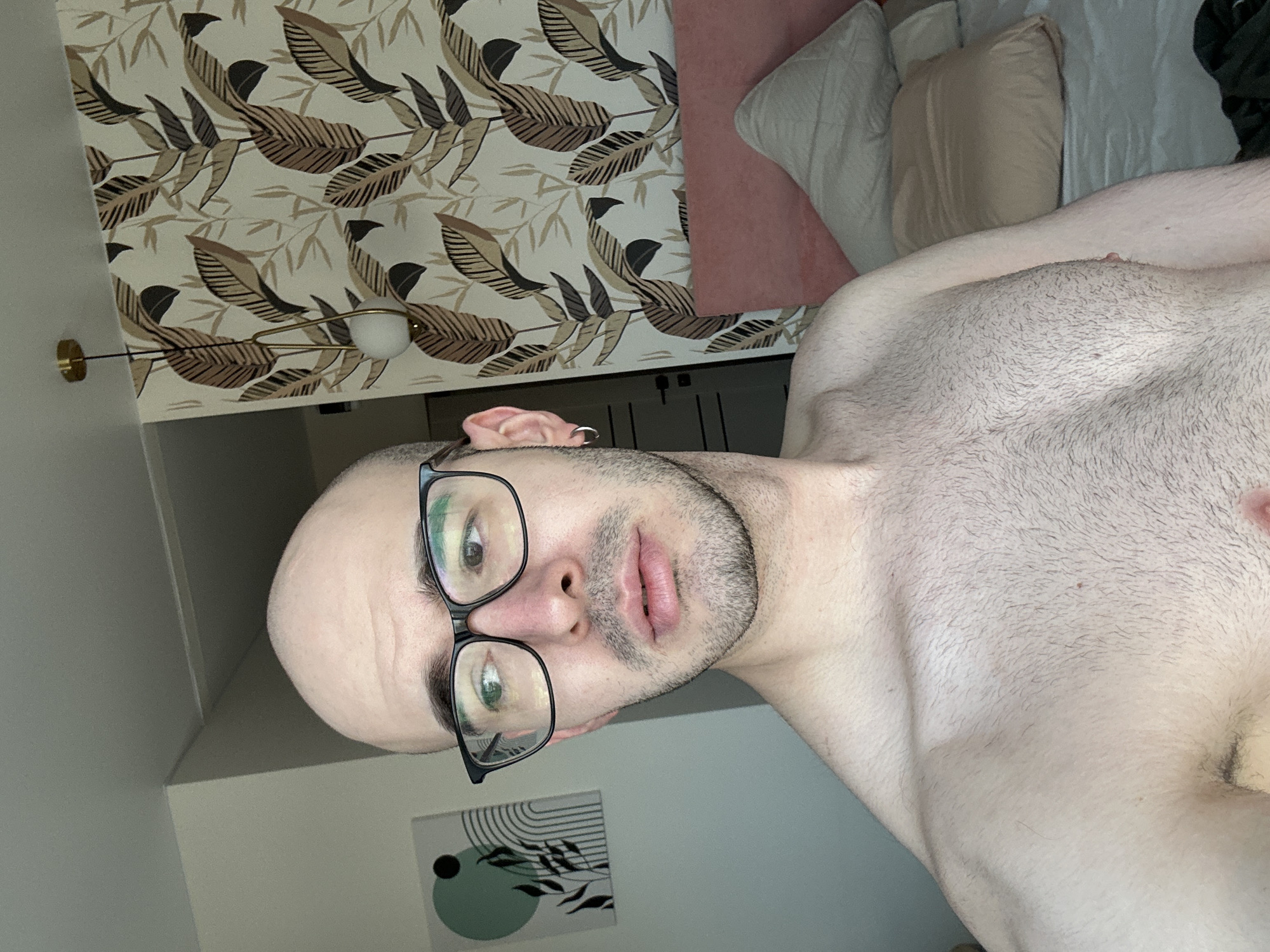 A close-up, angled self-portrait of a bald man with glasses and a short beard. He is shirtless and appears to be lying in bed. The background includes patterned wallpaper, pillows, a painting, and a lamp. There is no information to identify the location in the city of Kazan, Russia.