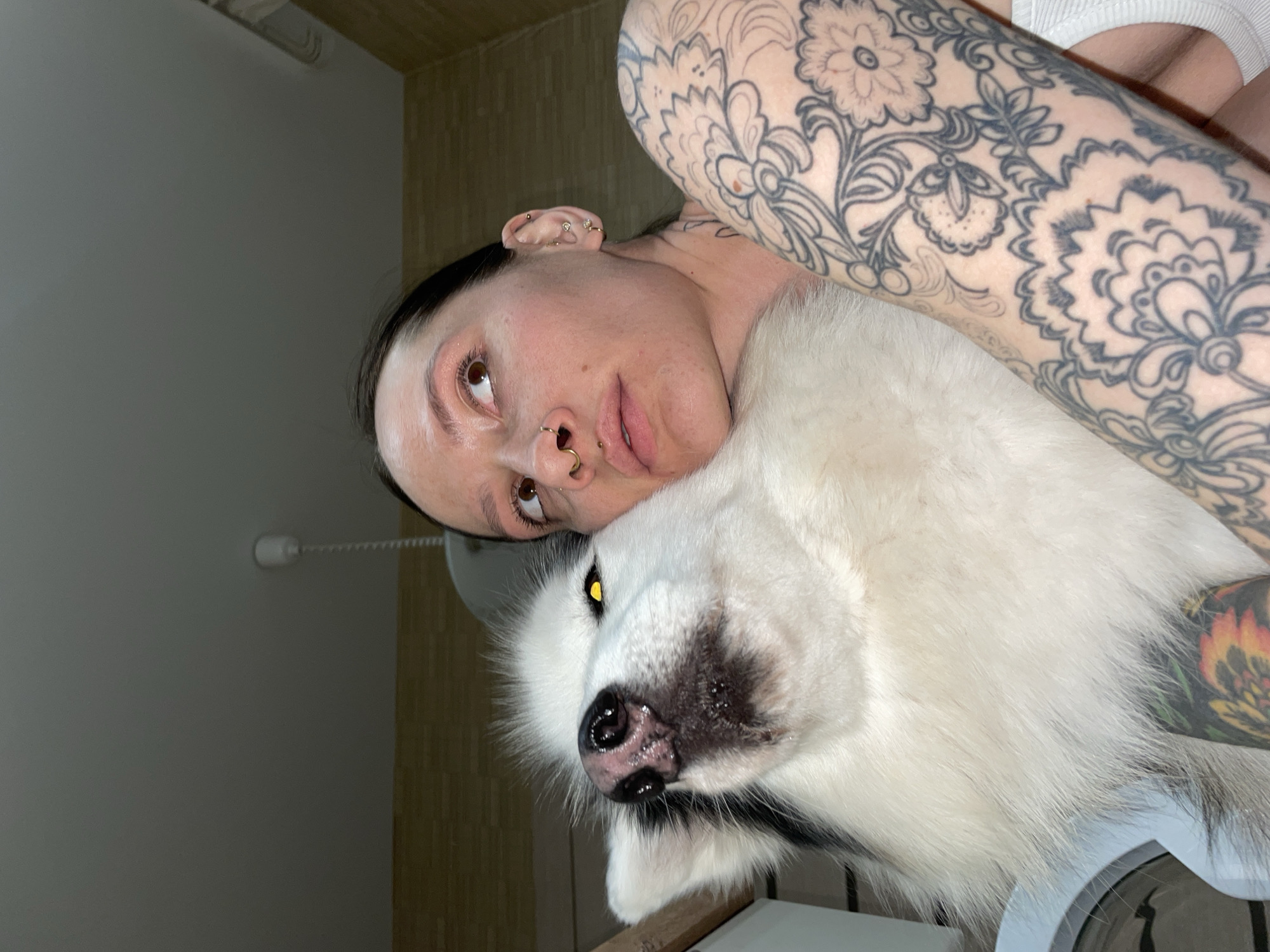 The scene depicts an indoor location in Izhevsk, Russia. A female individual is shown in close physical proximity to a large, fluffy dog. The female has dark hair, multiple facial piercings (septum ring, nostril ring), and several ear piercings on her right ear. Her right arm is extensively covered with black ink tattoos featuring floral and paisley patterns. She is wearing a white garment, visible on her right shoulder. Her face is pressed against the dog's head, and she gazes upwards and to the right with brown eyes. The dog, predominantly white with black markings, has a black nose and exhibits yellow eye shine in its visible left eye. Its head is tilted upwards, appearing to look towards the camera. The immediate background features a light grey ceiling with a white cylindrical fixture and a hanging cord, alongside a light-colored, textured wall.