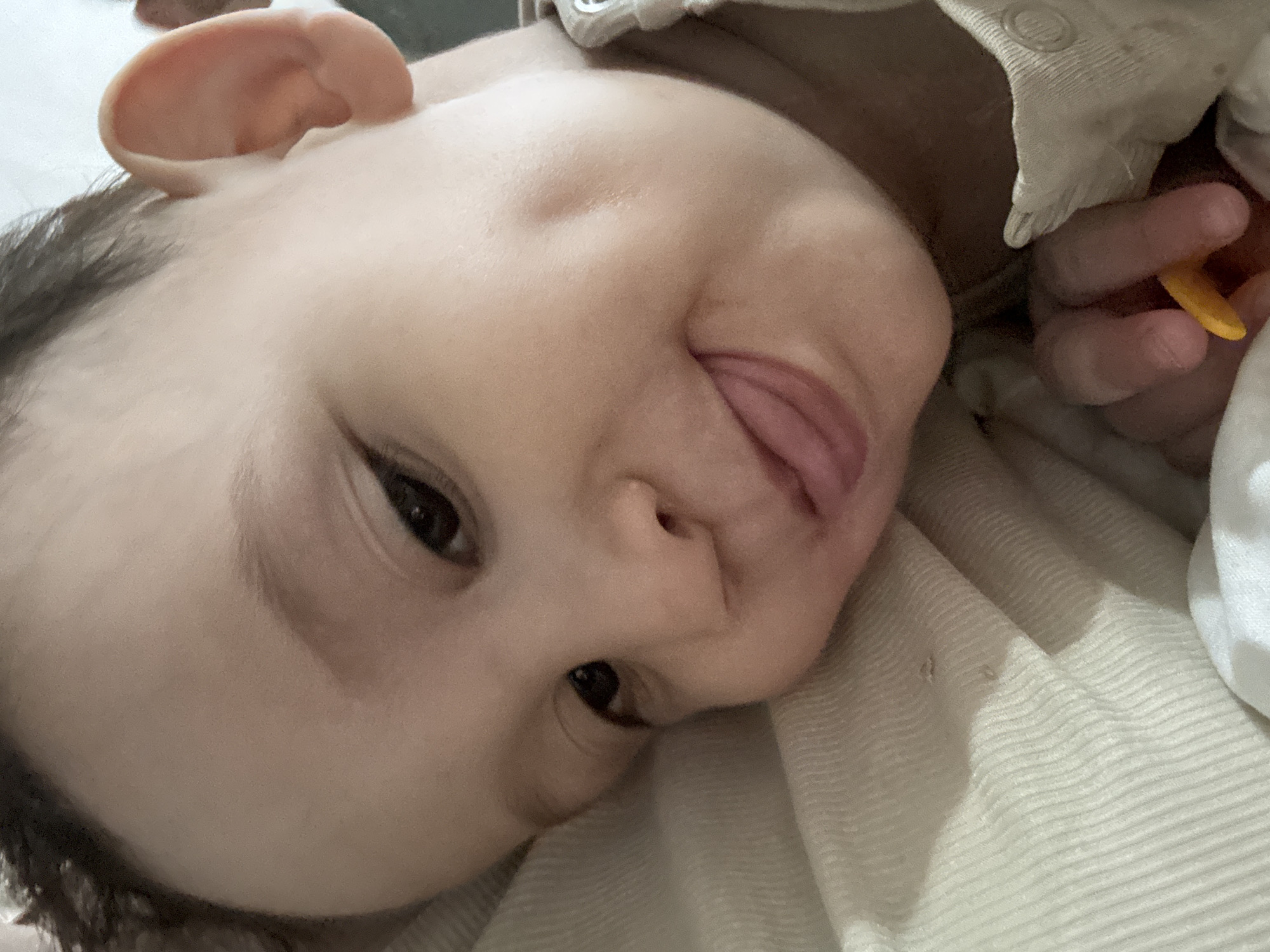 A close-up, angled view shows a baby lying down on a textured, light-colored surface, possibly a blanket or sheet. The baby's face is prominent, displaying its tongue sticking out. The baby has dark hair and eyes. A hand holding a small, yellow object is visible in the upper right corner. The photograph's location cannot be determined from the provided image.