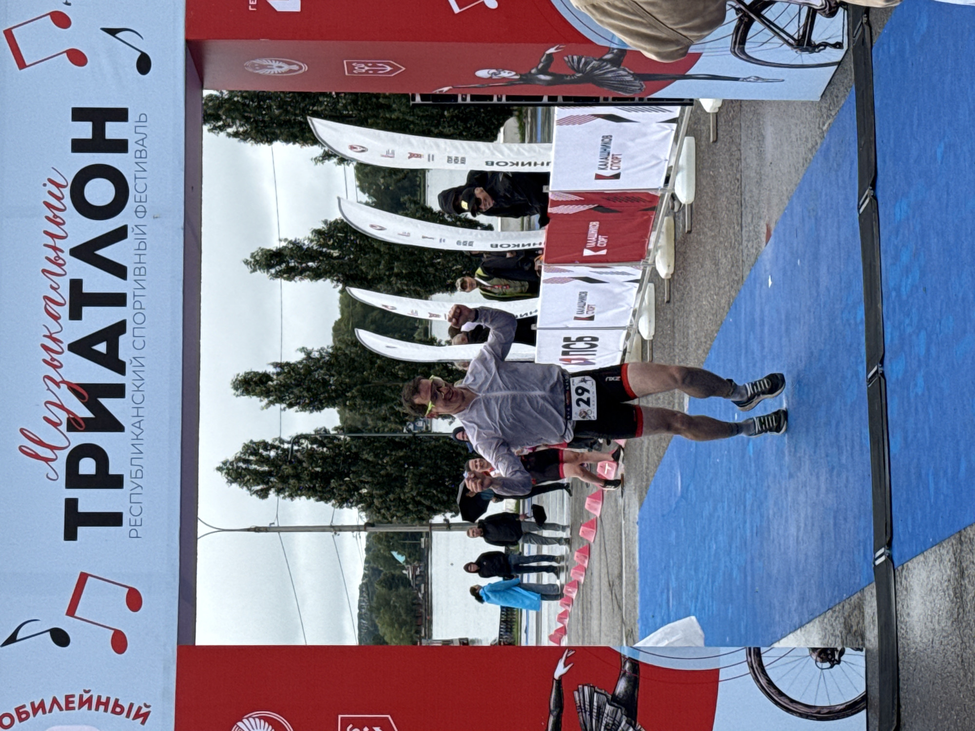A male athlete, wearing a grey, short-sleeved, zippered shirt, black cycling shorts with red trim, and grey athletic shoes, crosses a finish line. He has yellow-tinted goggles on his forehead and a bib number "29" on his shorts. The athlete appears to be celebrating, with his left arm raised in a fist and his mouth open in an excited expression. He is running on a blue mat that covers asphalt. In the background, a large red and white banner with musical notes and the text "Музыкальный ТРИАТЛОН" (Musical TRIATHLON) and "РЕСПУБЛИКАНСКИЙ СПОРТИВНЫЙ ФЕСТИВАЛЬ" (REPUBLICAN SPORTS FESTIVAL) is visible, suspended between two red archways. Behind the archways are trees, a body of water, and several spectators. To the right of the finish line, a red banner with a stylized depiction of a dancer and a bicycle wheel is present, along with other promotional banners. The event is identified as the "Musical Triathlon" in Votkinsk, Russia. The sky is overcast, suggesting a cloudy day.