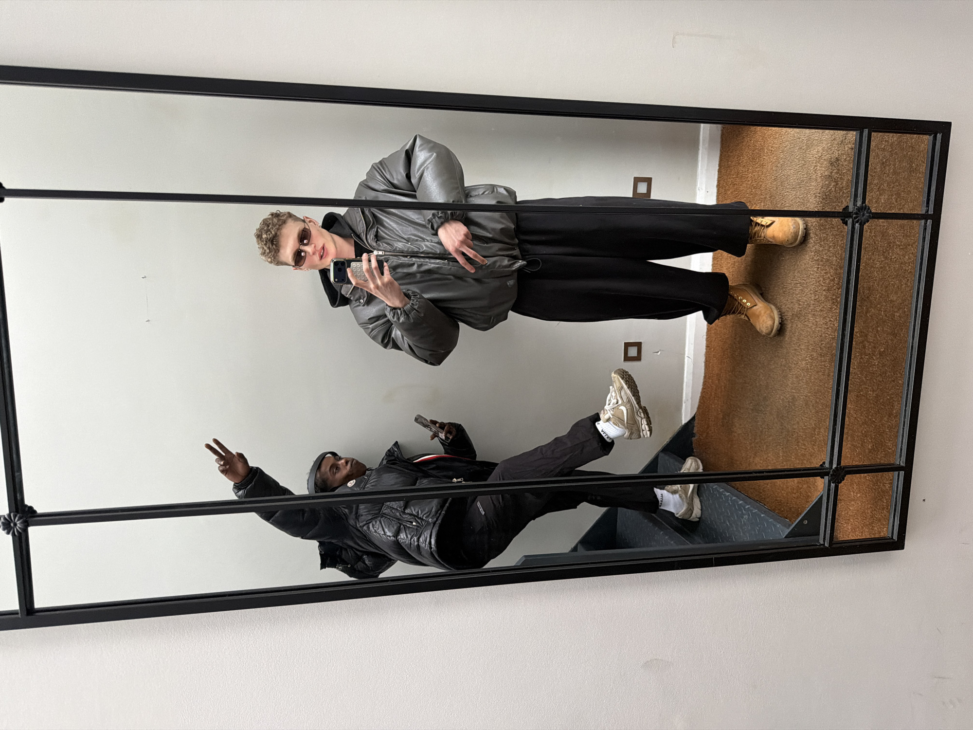 A large, rectangular, black-framed mirror reflects two people and their surroundings. The mirror frame has decorative spherical elements at its corners. The reflection shows two men in casual clothing. One man, positioned towards the top, is wearing a grey puffer jacket, black pants, sunglasses, and tan boots. He is holding a mobile phone and appears to be taking a photograph of himself. The second man, positioned towards the bottom, is wearing a black puffer jacket, black pants, a beanie, and off-white sneakers. He is holding a mobile phone and making a peace sign with his hand. The reflection reveals the surrounding environment. A plain, light-colored wall is visible behind both men. Above the first man, a section of brown carpet is reflected. Below the second man, grey stairs are visible. The absence of specific landmarks or street signs makes it impossible to determine the exact location in London, United Kingdom from the image alone.