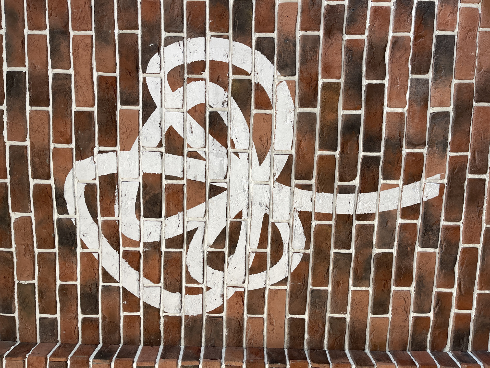 The image shows a brick wall with white graffiti. The wall is made of red and brown bricks with white grout. The graffiti is a white, looping design that resembles a knot or symbol. The location is Khimki, Russia.