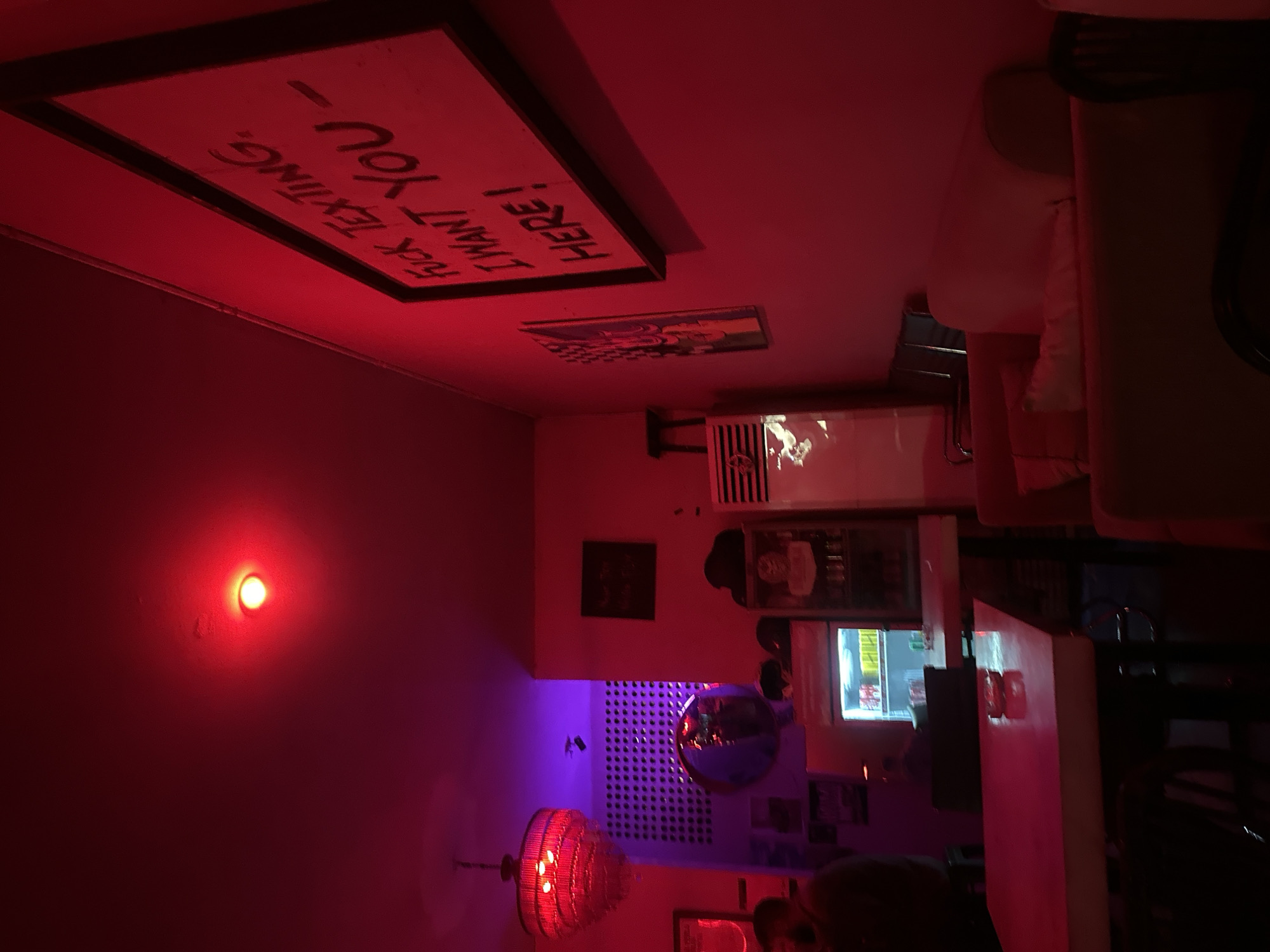 The image depicts the interior of a bar in North Kuta, Indonesia. The scene is dimly lit with red and purple lighting, creating an intimate atmosphere. A sign on the ceiling reads "HERE! I WANT YOU - F*CK TEXTING". A red light is affixed to a wall. Below the sign, a bar area is visible, featuring a counter with what appears to be bottles on top. A refrigeration unit is positioned against a wall. In the background, a decorative round mirror hangs on the wall along with other framed items. A red lamp hangs from the ceiling. The background features a display screen of sorts. Seating is present. An individual's head is seen in the lower portion of the image.
