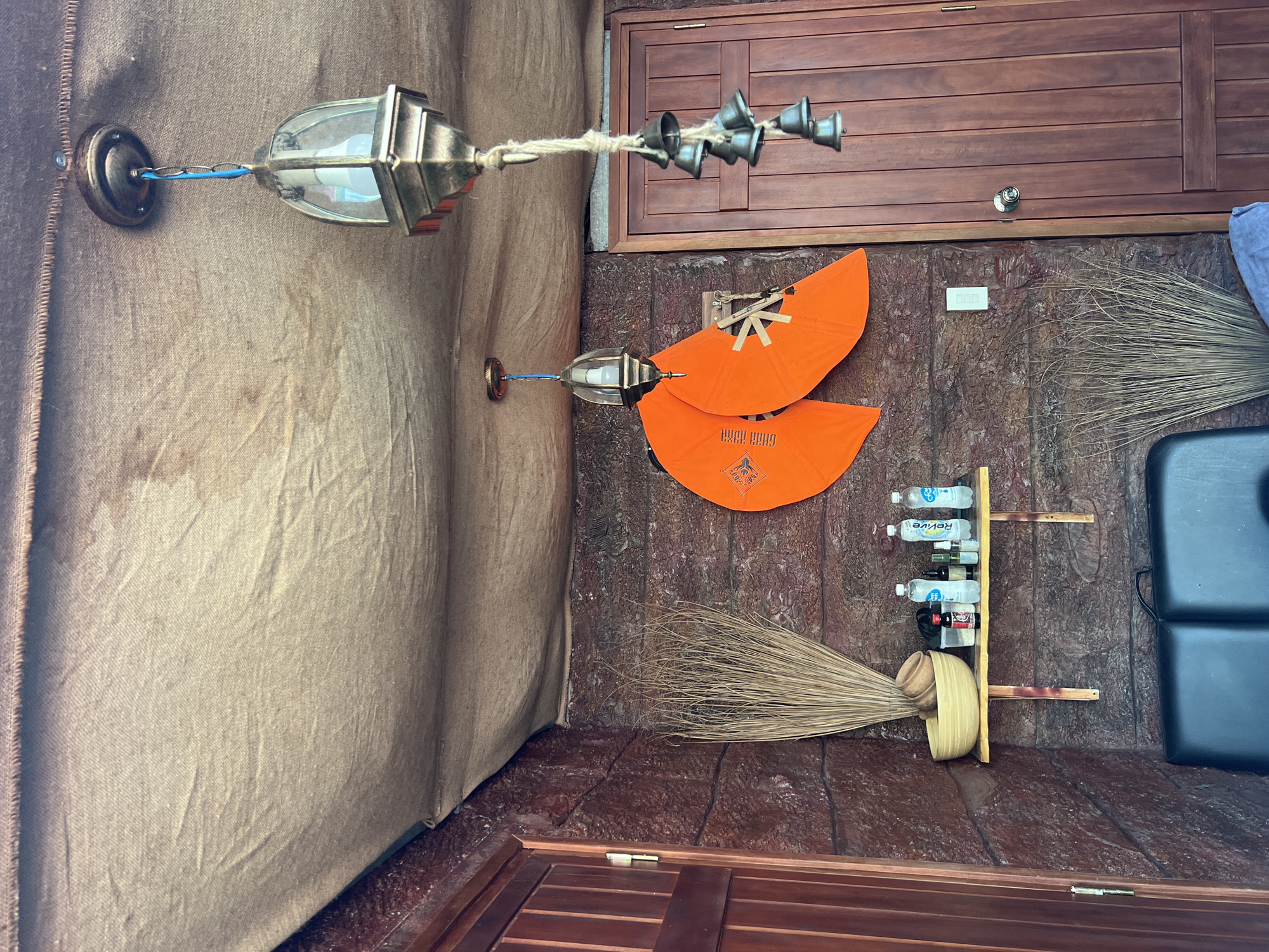 A rustic interior scene in what appears to be the Bac Natural Heritage Area Trang, Vietnam, features a corner of a room with walls covered in textured brown fabric and wood paneling. Two decorative metal lamps hang from the wall, one with a string of small bells. Below, two orange, fan-shaped decorations are mounted on the wall. Two brooms lean against the wall. A small rack holds bottles of water and wine, and a black rectangular object, likely a cushion or piece of equipment, is visible in the corner.