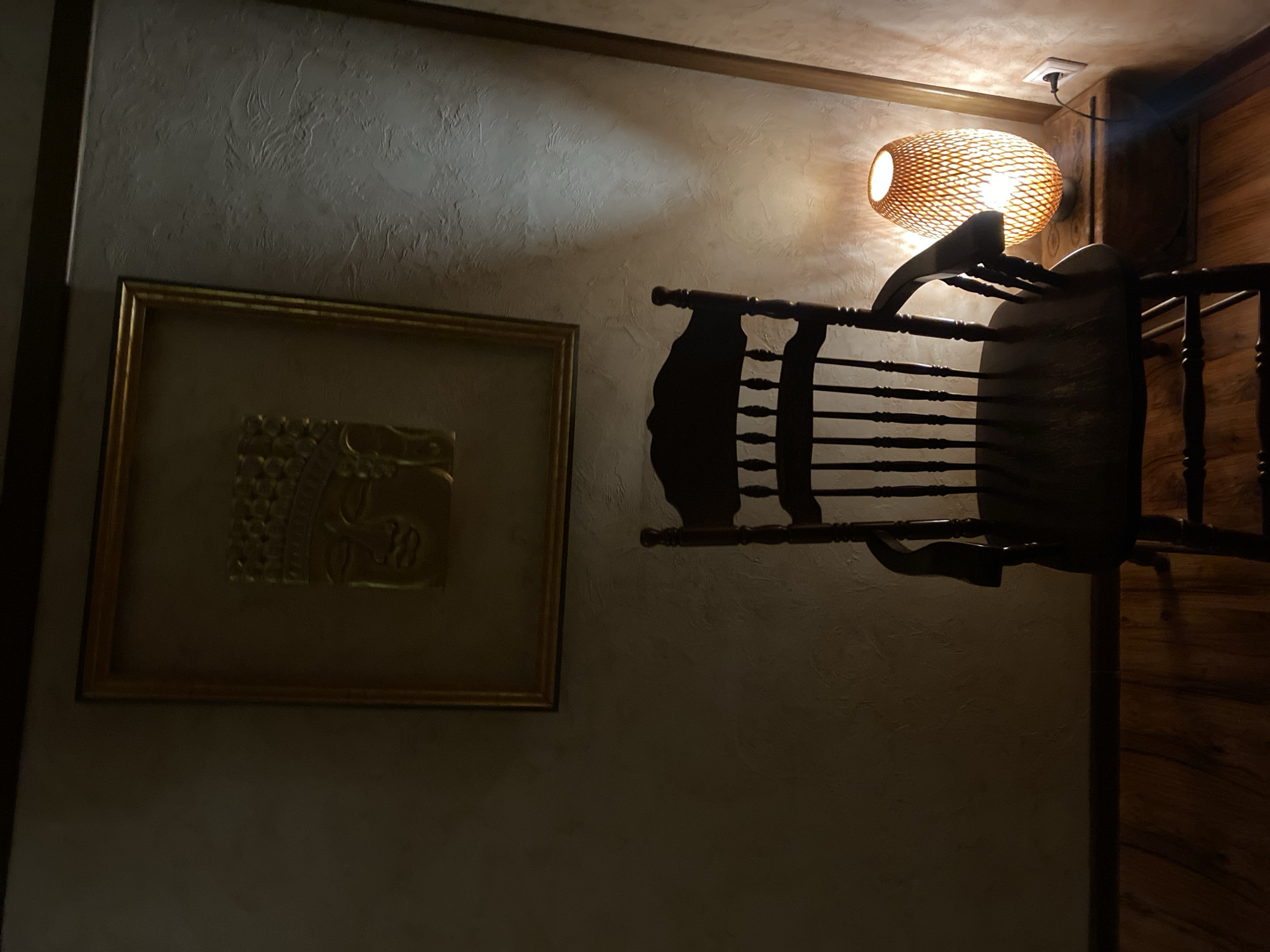 A dark wooden rocking chair is positioned in front of a textured wall, with a framed golden relief of a Buddha's face mounted on the wall above and slightly to the left of the chair. To the right of the chair, a woven, oval-shaped lamp emits a warm orange glow, illuminating a small wooden side table and the wall behind it. The floor is made of polished wooden planks. The scene is dimly lit, creating a serene and contemplative atmosphere. The location is Unknown, Unknown.