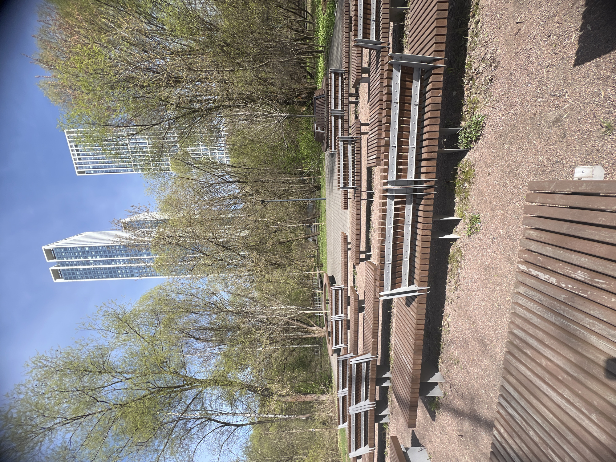 This image captures an outdoor urban park or green space in Khimki, Russia, on a bright and sunny day, likely in springtime, given the emerging green leaves on some trees. The scene is dominated by a series of modern, tiered wooden benches with silver-colored metal supports, arranged in a structured, amphitheater-like fashion along a gravel path. A paved walkway runs through the park further back from the benches.
No people are visible, suggesting the area is currently unoccupied and quiet. The strong sunlight casts clear shadows, indicating it is late morning or early afternoon. In the middle ground, numerous deciduous trees, some beginning to leaf out with light green foliage and others still bare, line the paths. A tall, slender light post stands among the trees.
The background features several modern, tall high-rise buildings with many windows, reflecting contemporary urban architecture, set against a clear blue sky. The overall impression is of a well-maintained, functional, and aesthetically pleasing urban leisure area. No text is visible in the image.