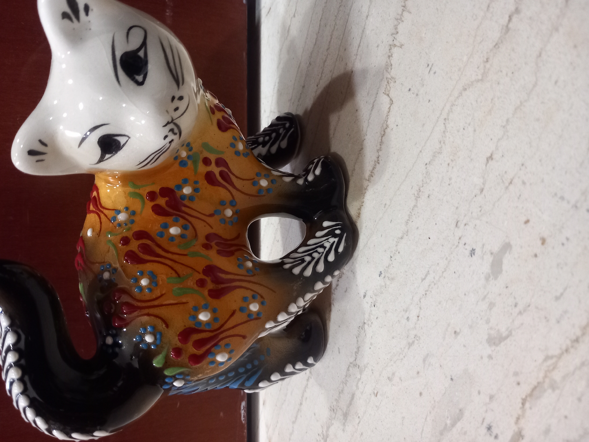 A close-up shot features a ceramic figurine of a cat with a white head and torso, adorned with vibrant patterns. The cat's face is depicted with black painted eyes and whiskers. The body is primarily orange, decorated with a repeating pattern of red and blue floral designs, along with swirling red motifs. Its tail and legs are black, detailed with white decorative elements. The figurine rests on a textured, light-colored surface, possibly marble or a similar stone, which appears slightly worn. A shadow is cast on the surface to the right of the figurine, suggesting the presence of a light source. The background on the left is dark and out of focus, hinting at a wooden surface or furniture. The lighting is dim, creating a moody atmosphere. There is no visible text in the image.