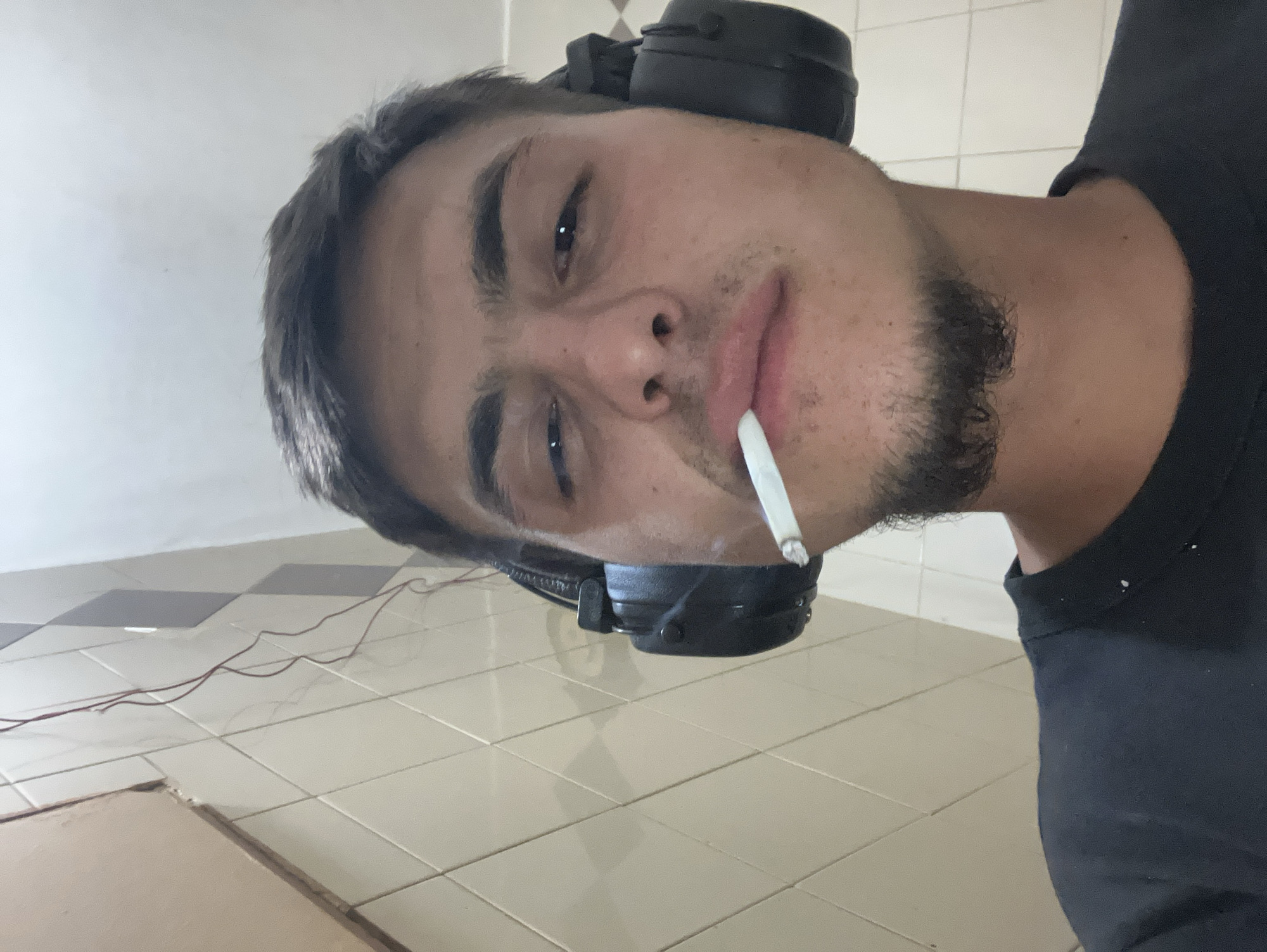 A selfie taken in an interior space. The subject is a young man with short dark hair, dark eyebrows, and stubble. He is wearing black headphones over his head and a black t-shirt. He holds a lit cigarette in his mouth. The background shows a tiled floor and part of a wall. There's a wire or cable visible on the floor. Location cannot be determined from image.