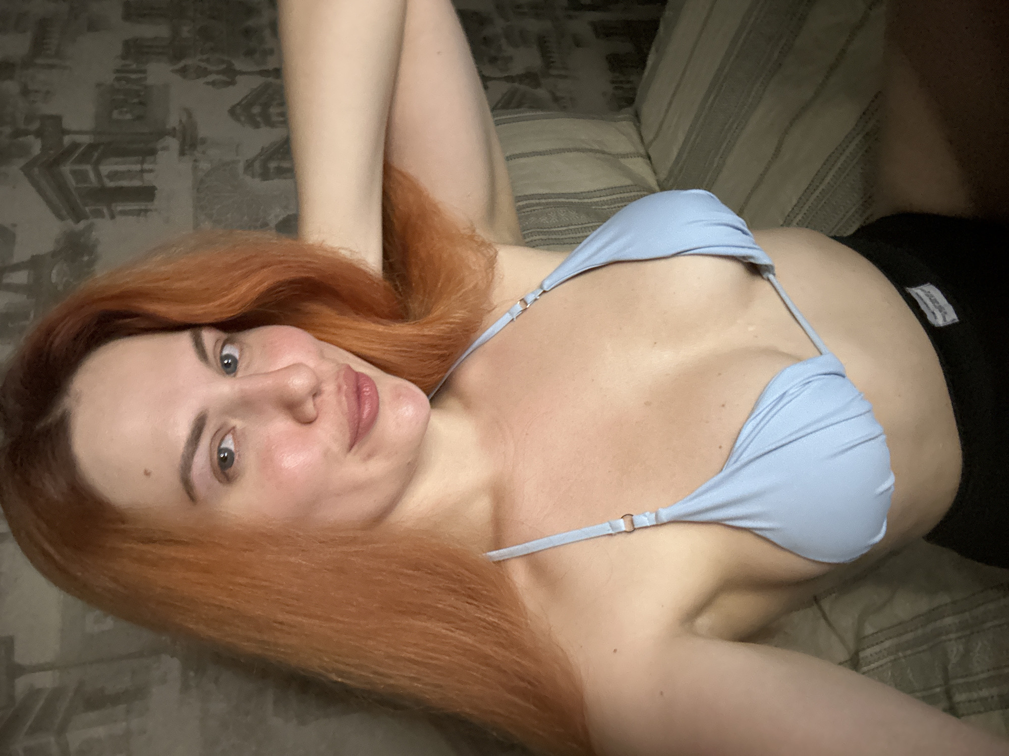 A young woman with long, auburn hair and fair skin is posing for a selfie. She is wearing a light blue bikini top and black shorts. Her right arm is raised and bent, with her hand resting on her head. She is looking at the camera with a slight smile. The background is a wall with a patterned wallpaper depicting Parisian landmarks. The lighting is dim, suggesting the photo was taken indoors.