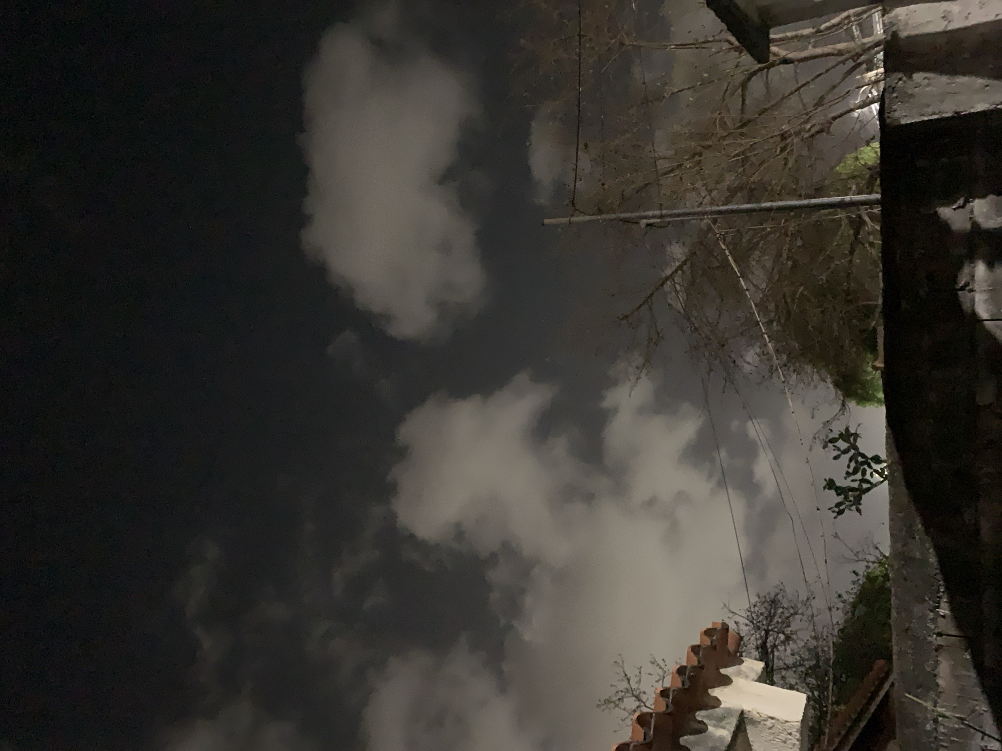 The image, taken at night, presents an upward-angled view of a sky partially obscured by clouds. In the foreground, architectural elements of a building are visible, featuring terracotta roofing tiles and a white stone structure. To the right, a dark, textured wall rises, potentially a part of the same building. Bare tree branches extend into the frame, silhouetted against the night sky. Utility poles and wires are also apparent, intersecting the view of the sky and trees. The location is suggested to be Santa Ana, Venezuela.