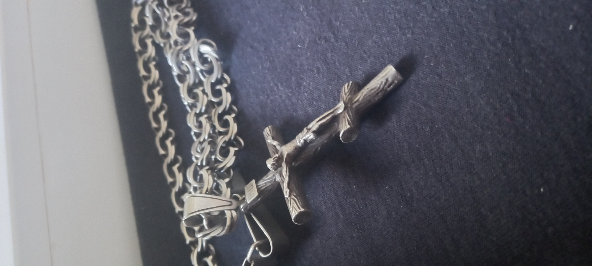 A close-up shot shows a silver metal cross pendant with a figure of Jesus affixed to a matching metal chain, resting on a dark blue or black fabric surface. The cross is designed to resemble rough wooden beams. The metal appears slightly tarnished. A white or light-colored vertical surface is visible on the far left edge of the frame. The location is unknown; the city of Kryvyi Rih, Ukraine cannot be determined from the image.
